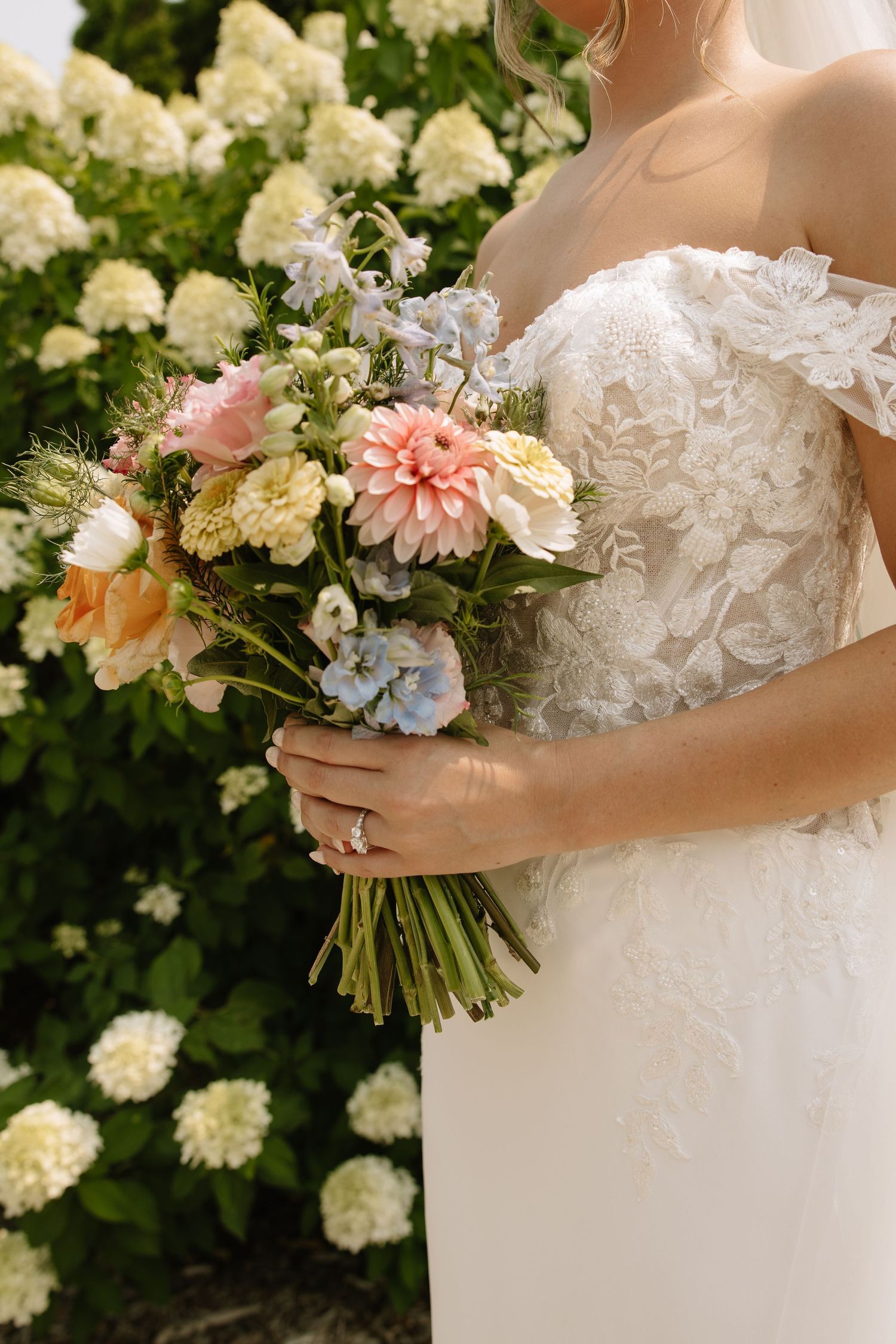 Bride in lace wedding gown holding a pastel bouquet of dahlias and hydrangeas in front of white blooming shrubs.