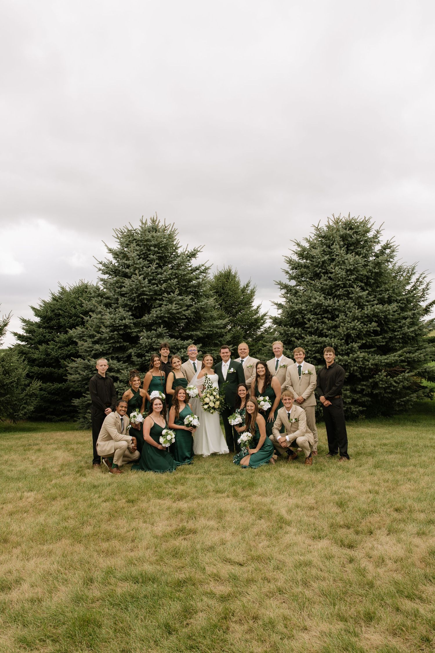 Wedding party poses outdoors in green and tan attire before evergreen trees on a cloudy day.
