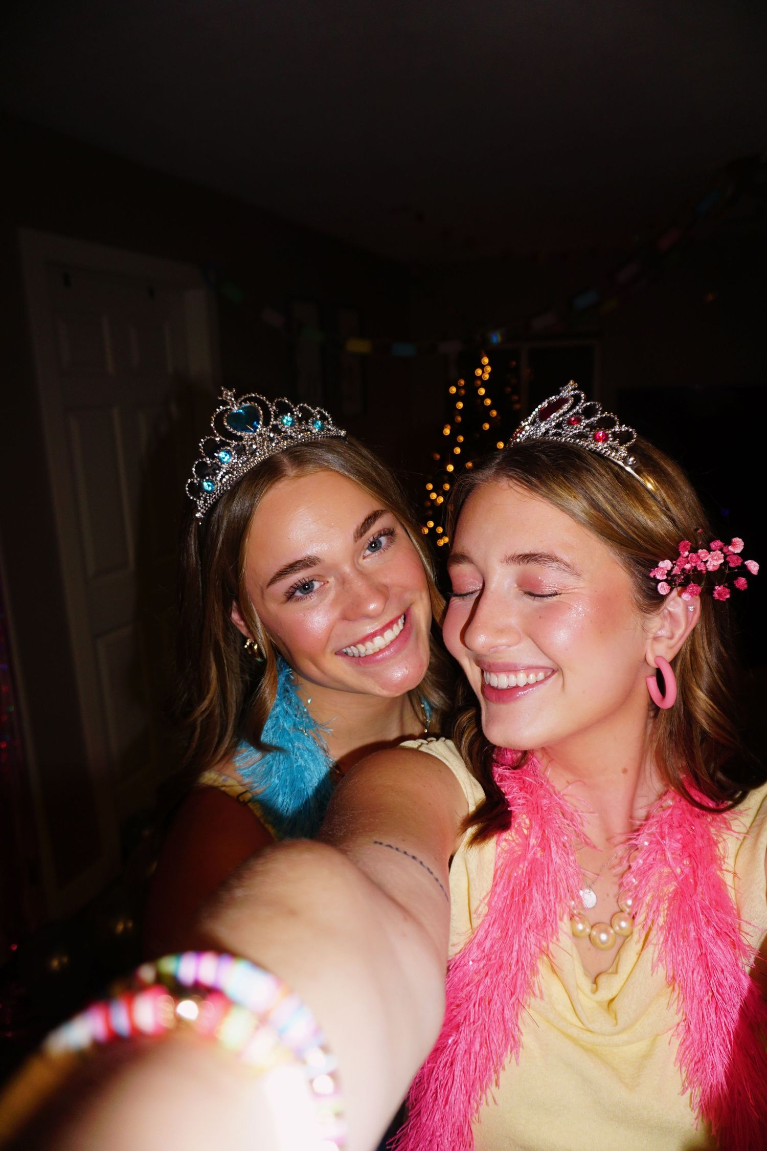 Two young women wearing tiaras and colorful accessories laughing together at a festive party.