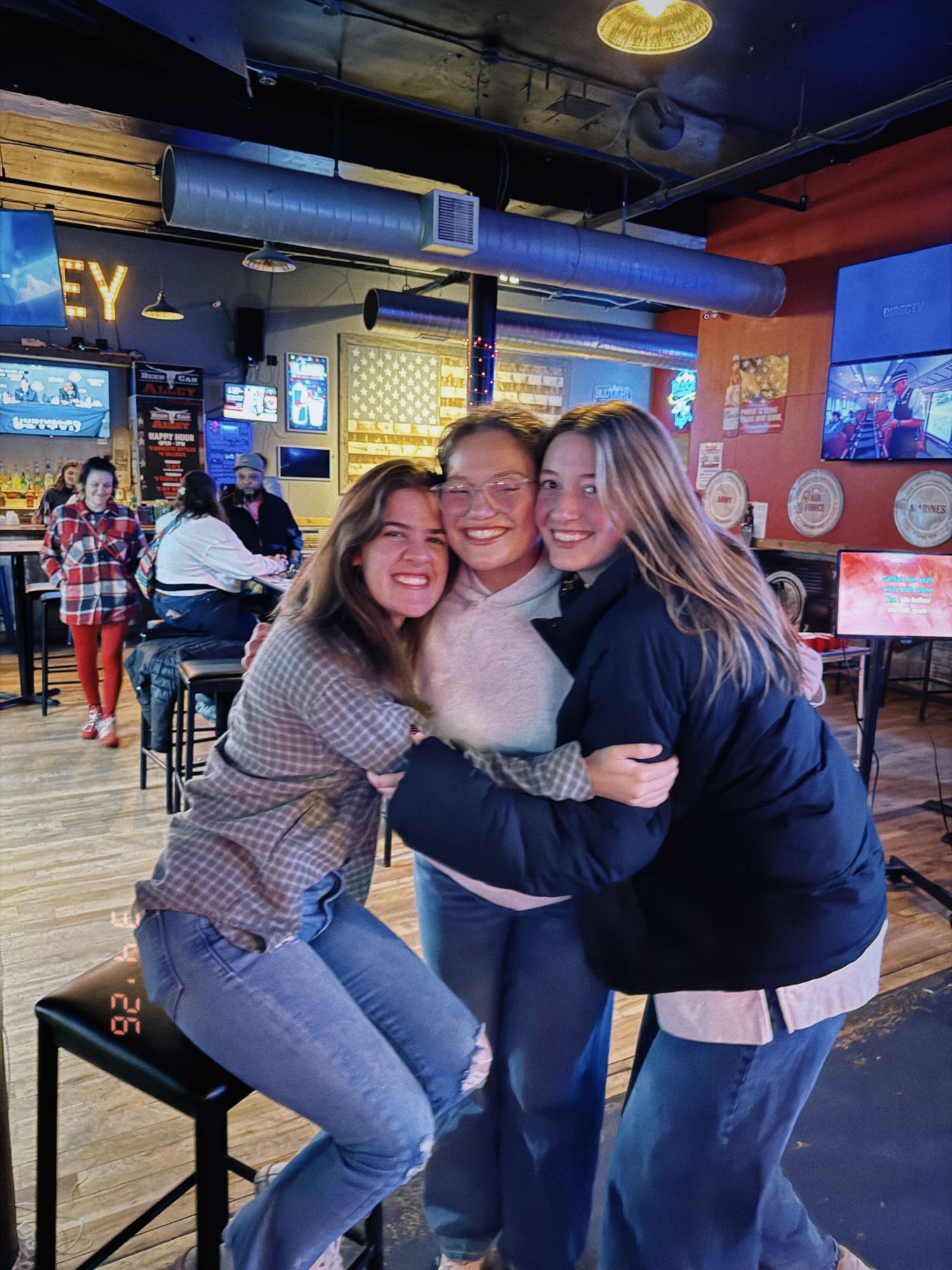 Three happy young women share a group hug at a casual restaurant bar setting.