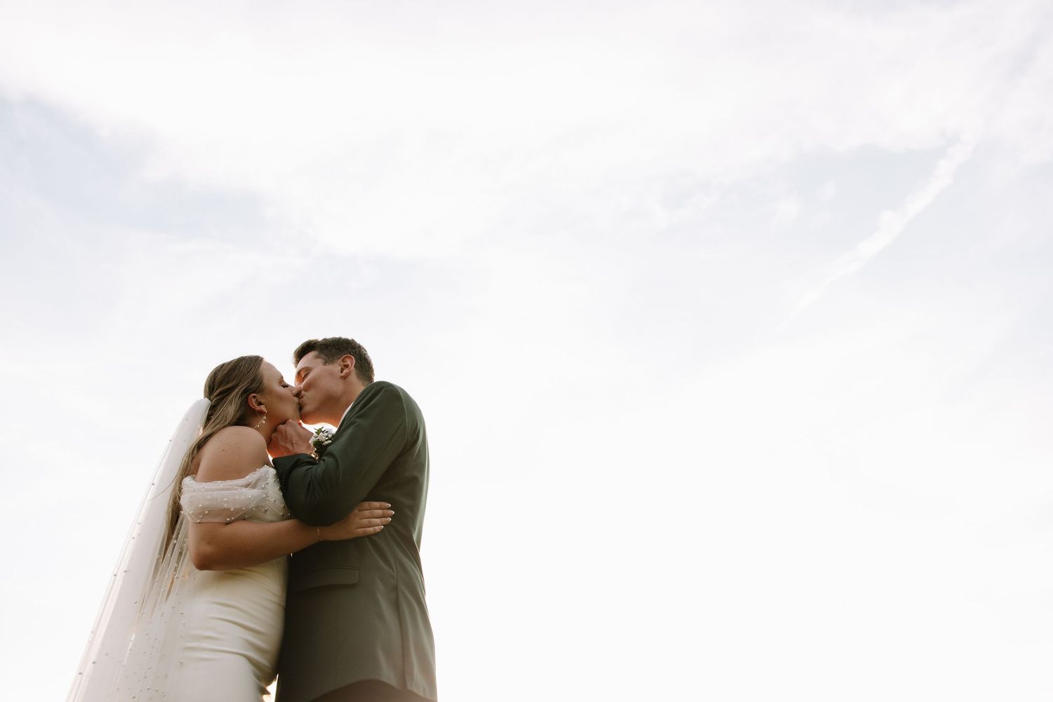 Bride and groom sharing a romantic kiss outdoors against a bright sky on their wedding day.