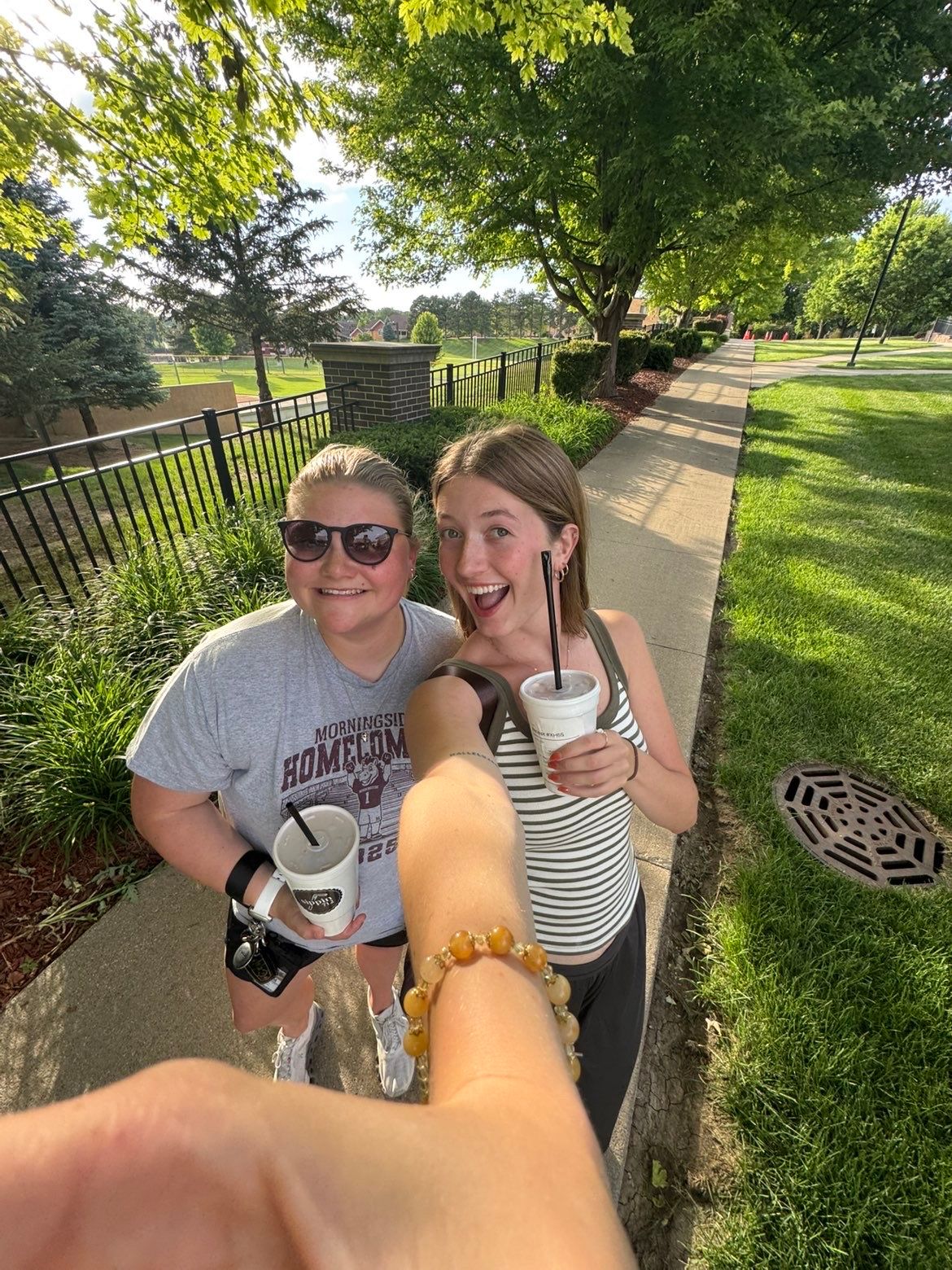 Two smiling young women pose on a sunny sidewalk holding drinks, surrounded by lush green trees and grass.