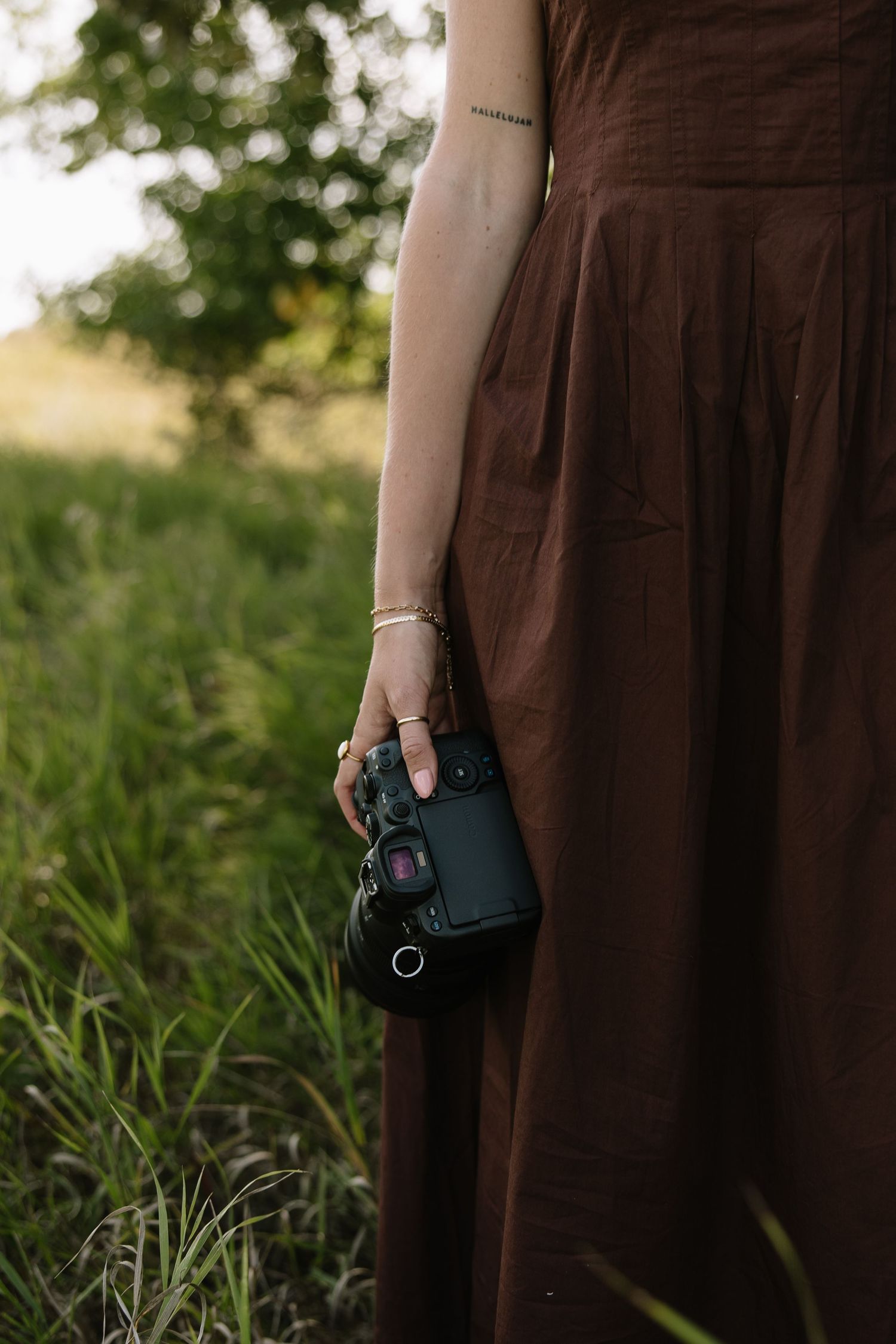 Woman in brown linen dress holding a DSLR camera while standing in a lush green field at golden hour.