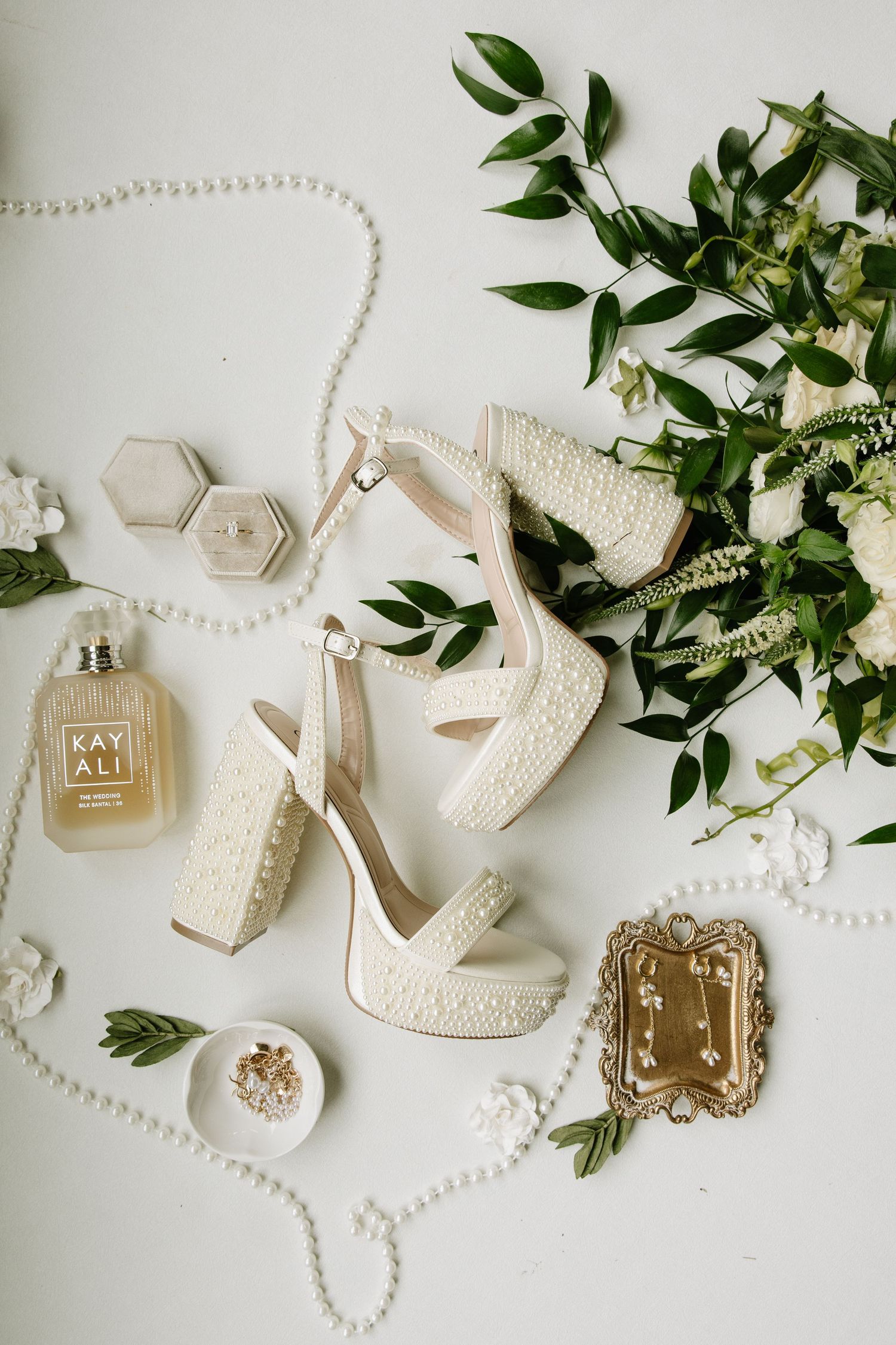 Pearl-adorned bridal heels surrounded by Kay Ali perfume, pearl necklace, ring box, and floral bouquet on white surface.