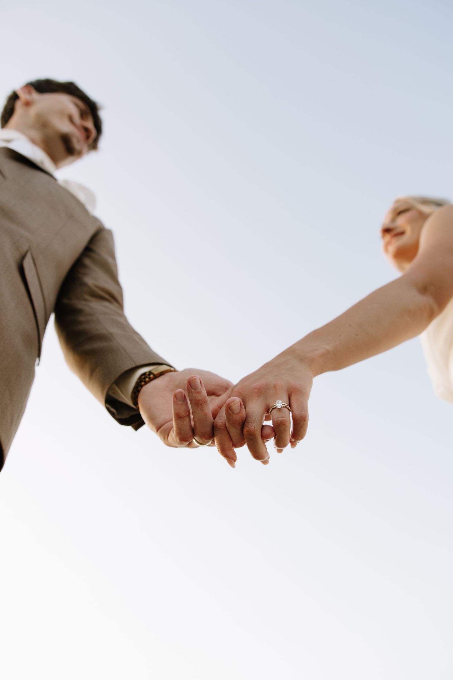 Bride and groom holding hands with wedding rings visible, shot from below against a clear sky.
