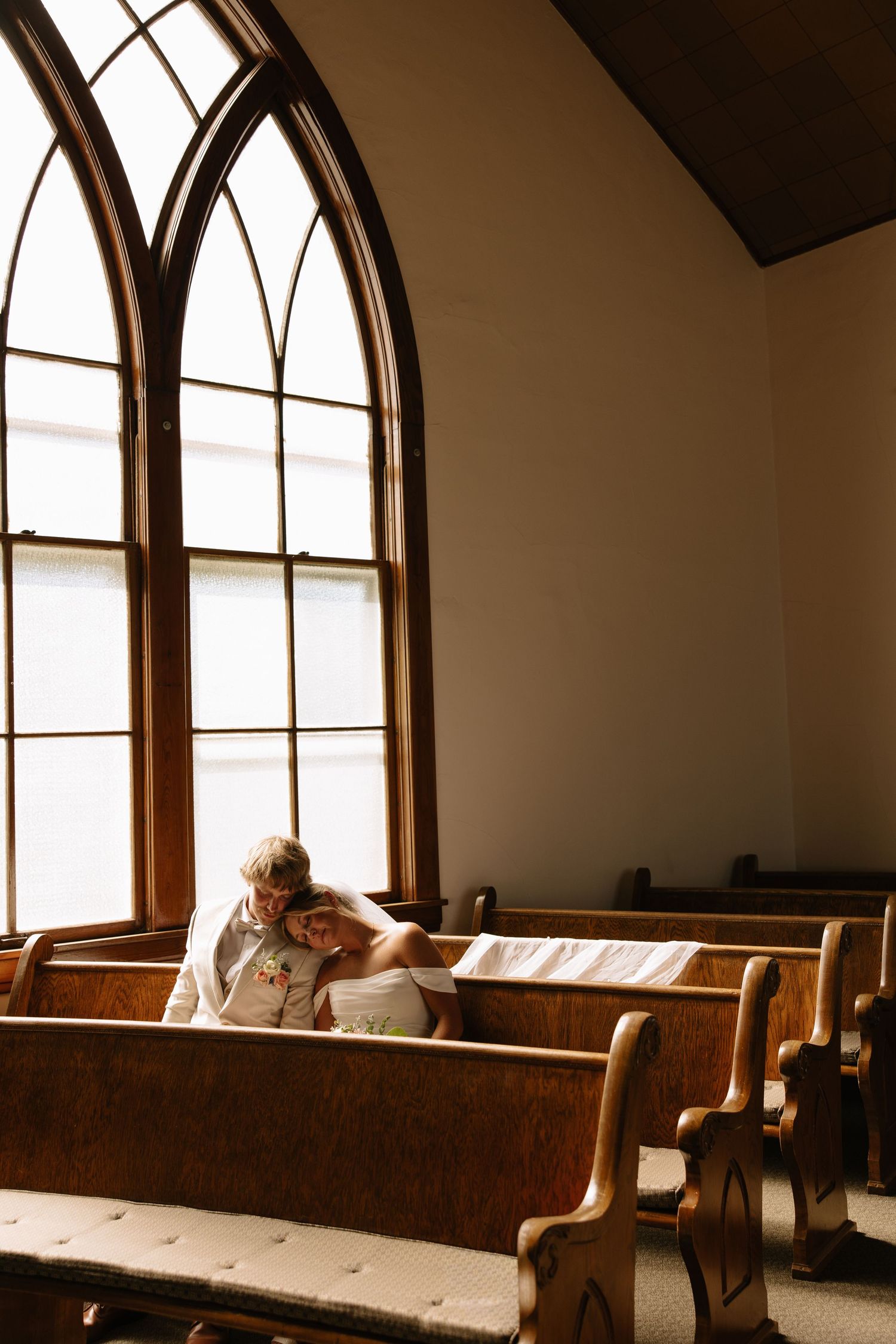 Couple sitting together in wooden church pews near a Gothic arched window, sharing an intimate moment.