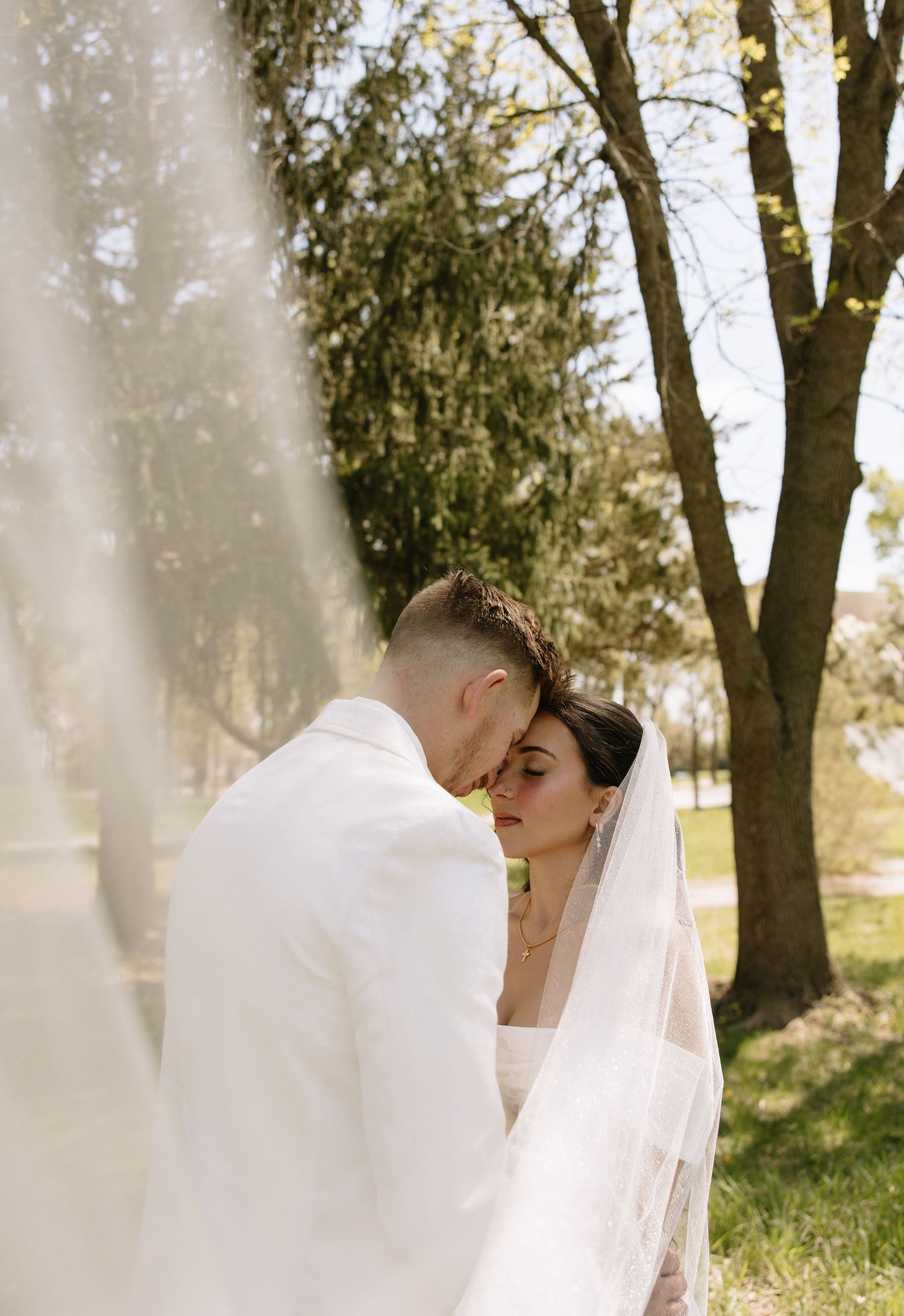 Bride and groom share an intimate forehead touch moment outdoors under trees with veil flowing.