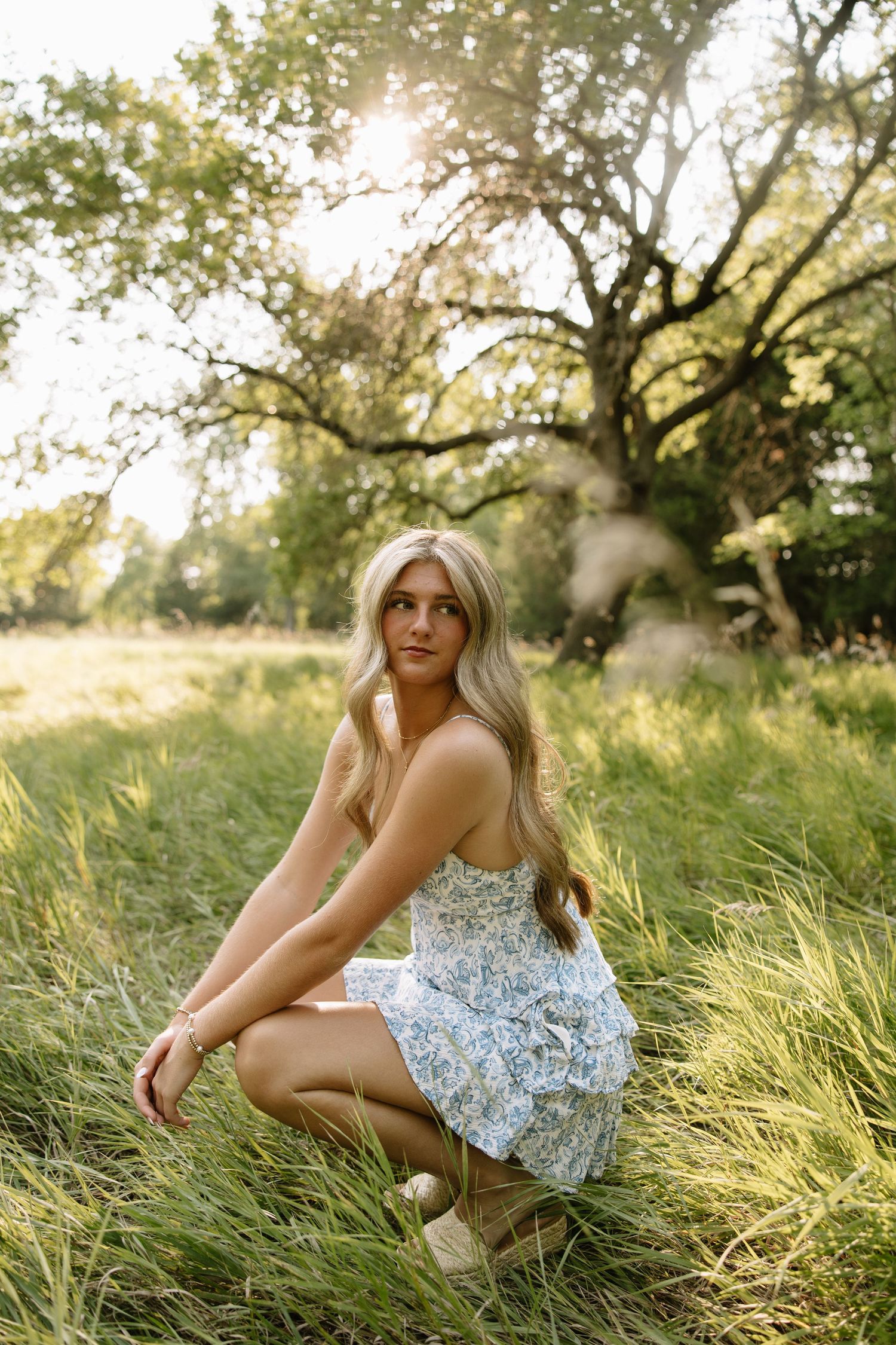 Blonde woman in floral sundress crouching in tall grass with sunlight filtering through trees in background.