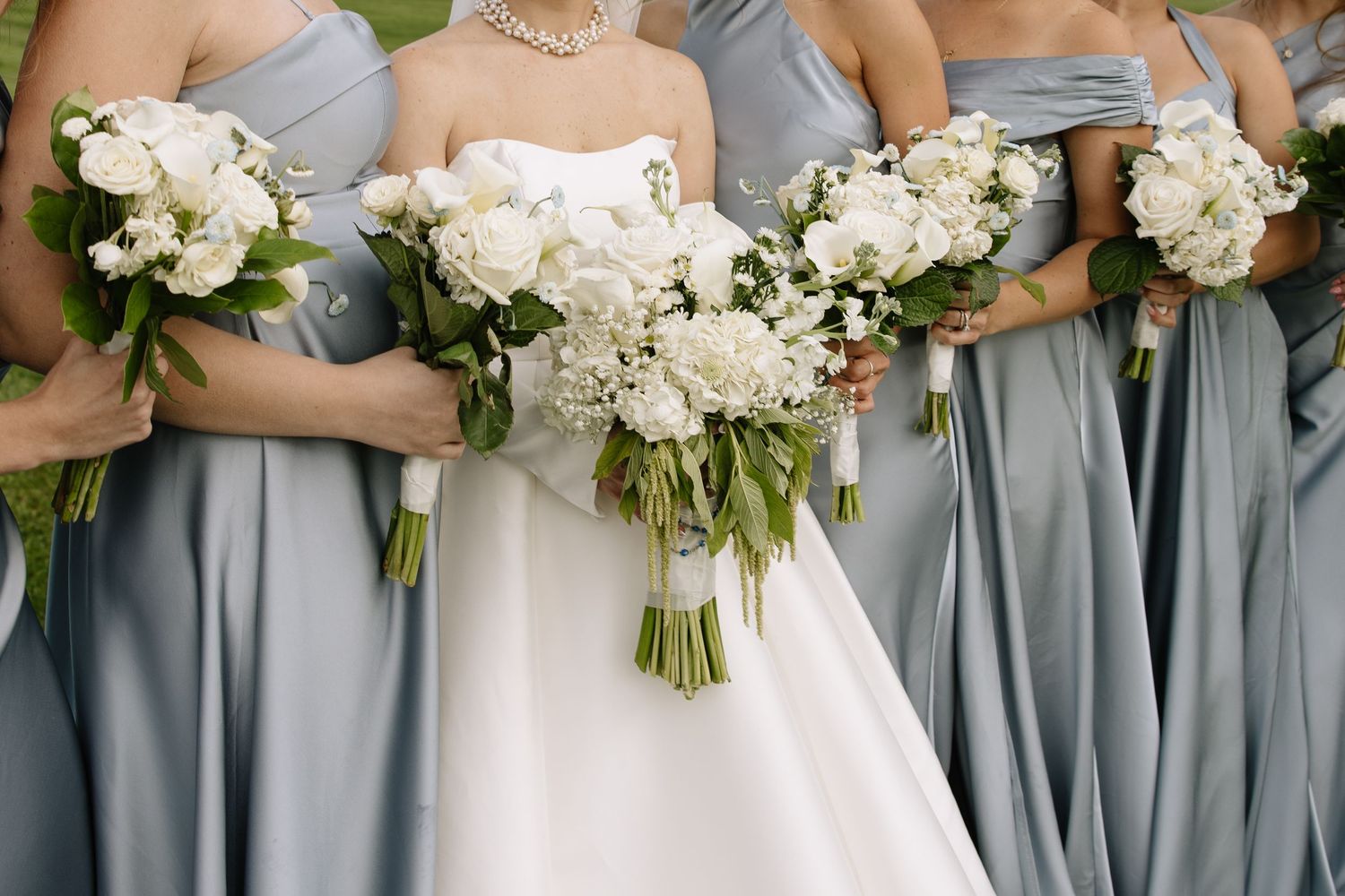 Bride and bridesmaids in dusty blue gowns holding elegant white floral bouquets with green foliage at a wedding.