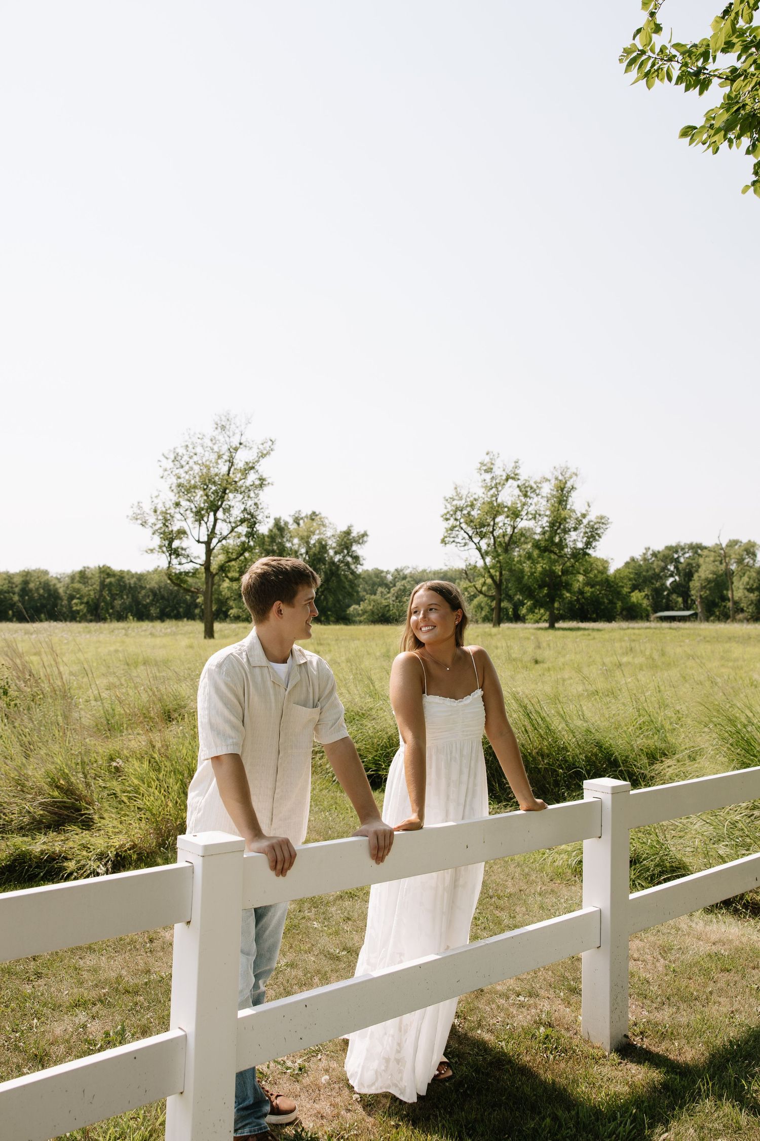 A couple in white outfits smiles at each other leaning on a white fence in a sunny green countryside.