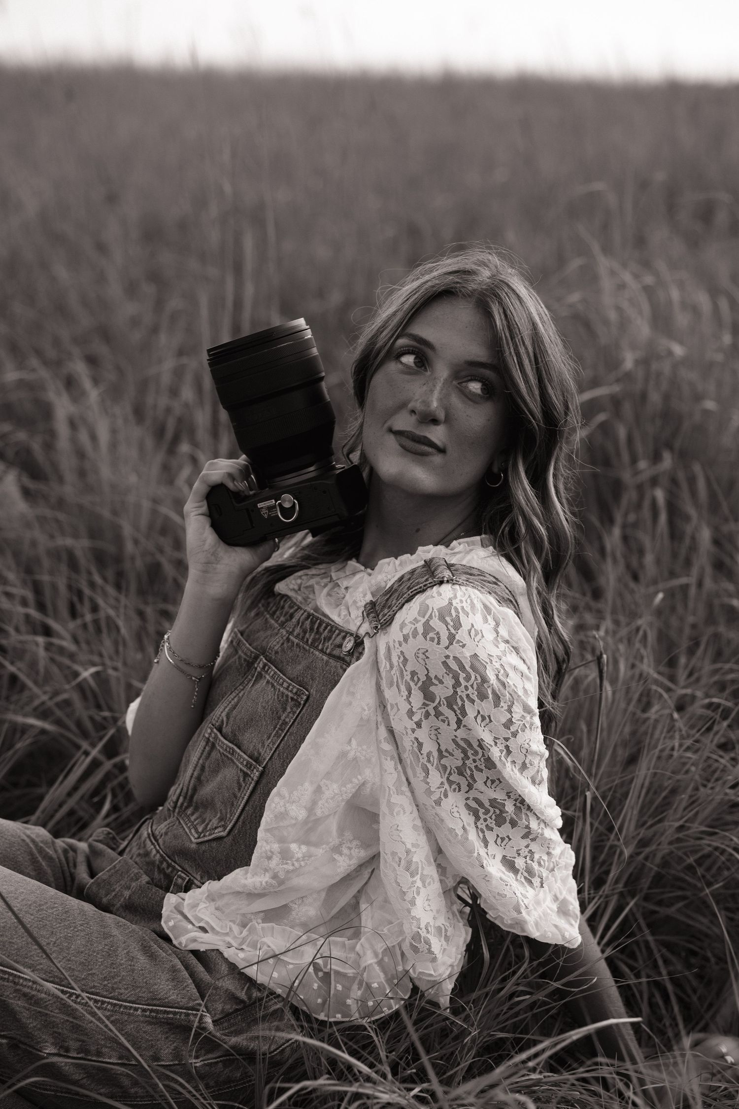 Young woman in denim overalls and lace top holds a camera in a tall grass field, black and white photo.