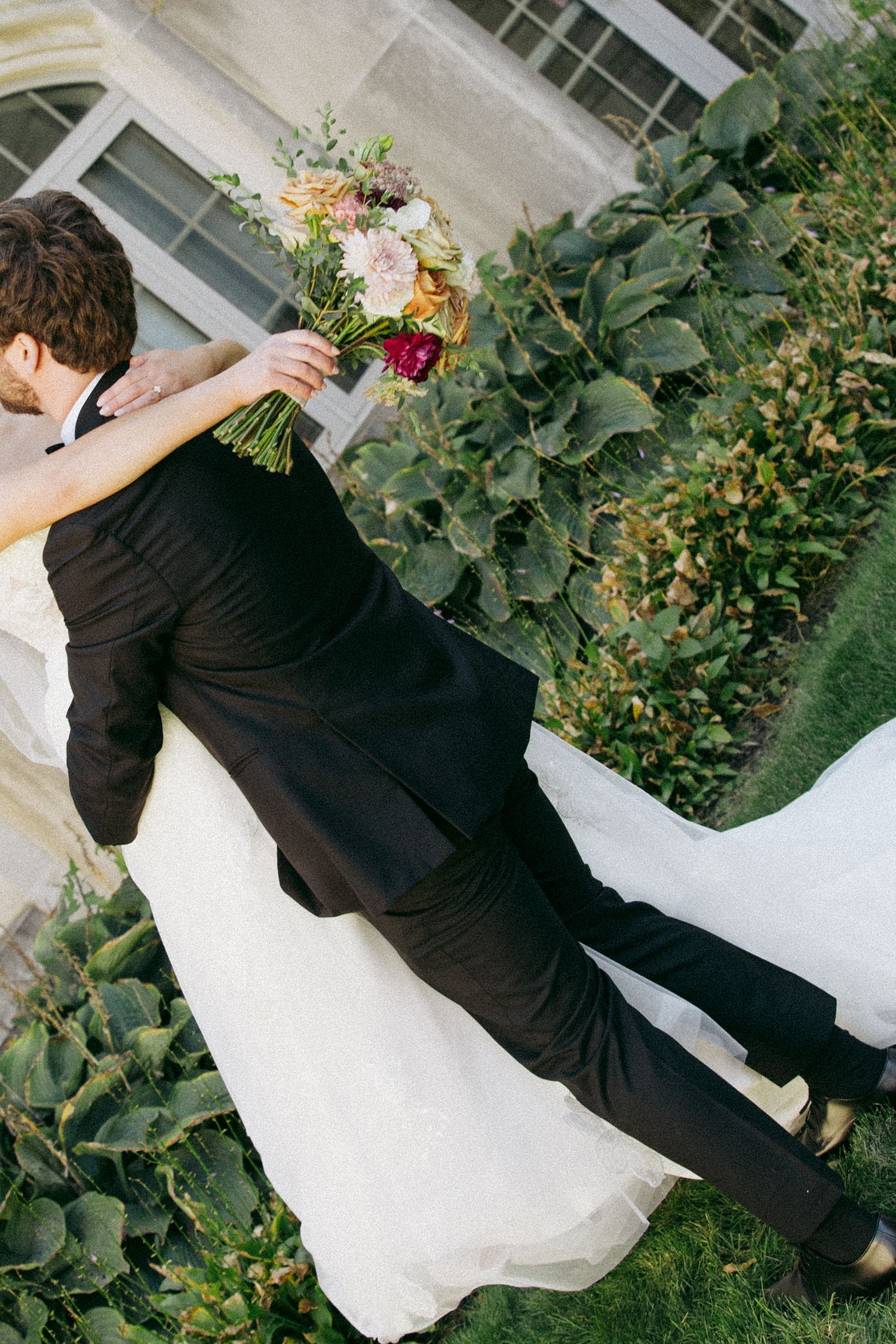 Bride in a white wedding gown embraces a groom in a black suit while holding a colorful bouquet beside lush green hostas.