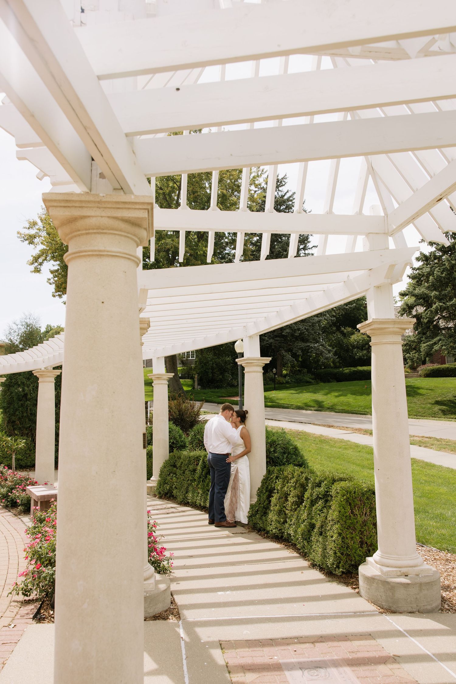 Couple sharing a romantic kiss under a white pergola surrounded by lush gardens on their wedding day.