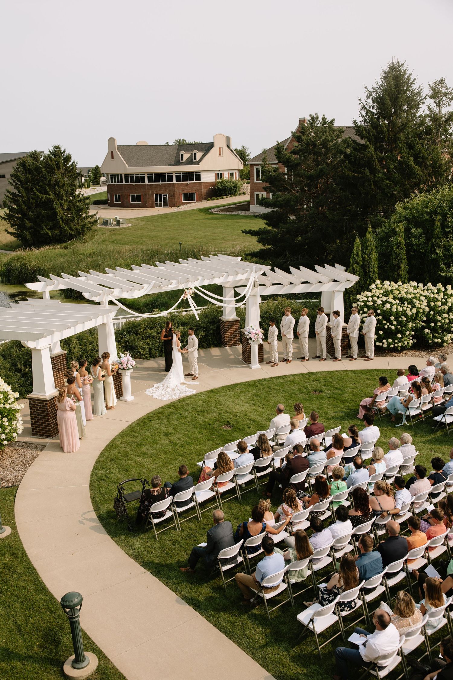 Aerial view of an elegant outdoor wedding ceremony on a manicured lawn with white pergola, bridal party, and seated guests.