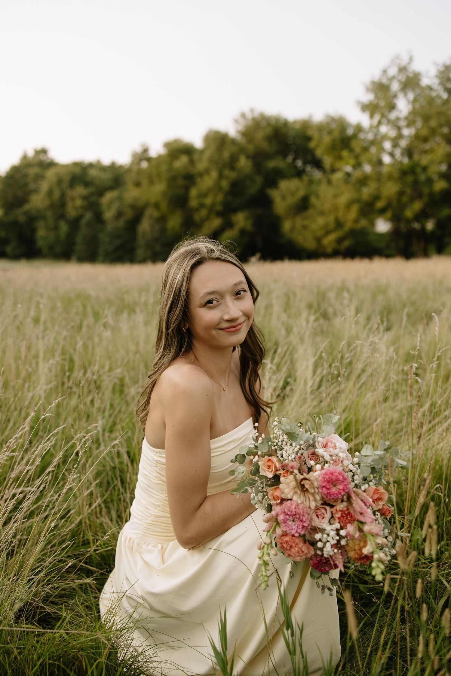 Bride in white strapless gown holds colorful bouquet while sitting in tall grass field with trees in background.