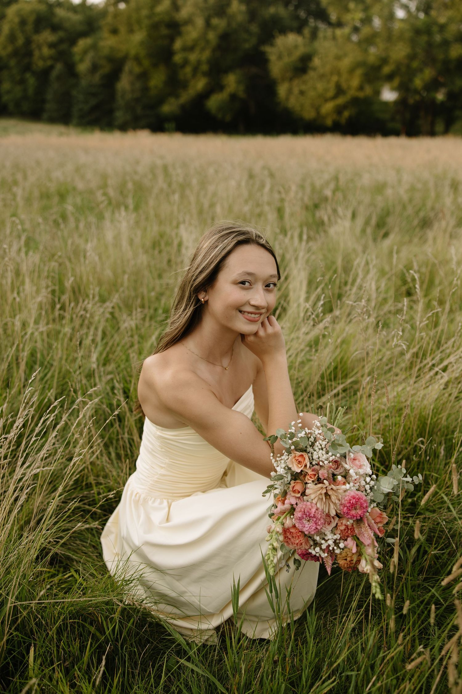 Smiling bride in white dress crouching in tall grass field holding colorful pink floral bouquet outdoors.
