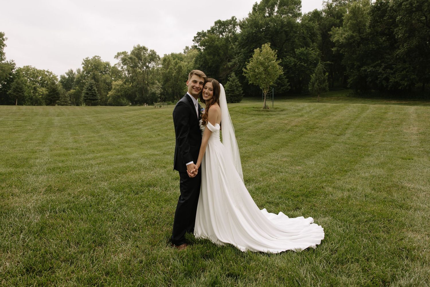 Bride and groom posing together on lush green lawn, bride wearing flowing white gown with long veil.