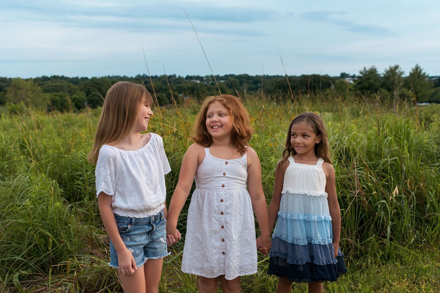 Three girls in white and blue outfits hold hands smiling in a lush green meadow under a cloudy sky.