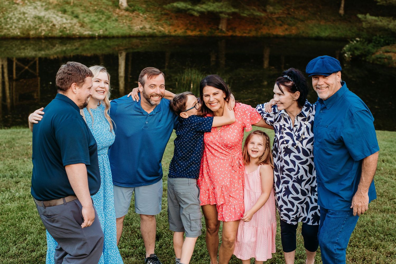 Happy family group of seven people smiling and embracing outdoors near a pond on a sunny day.