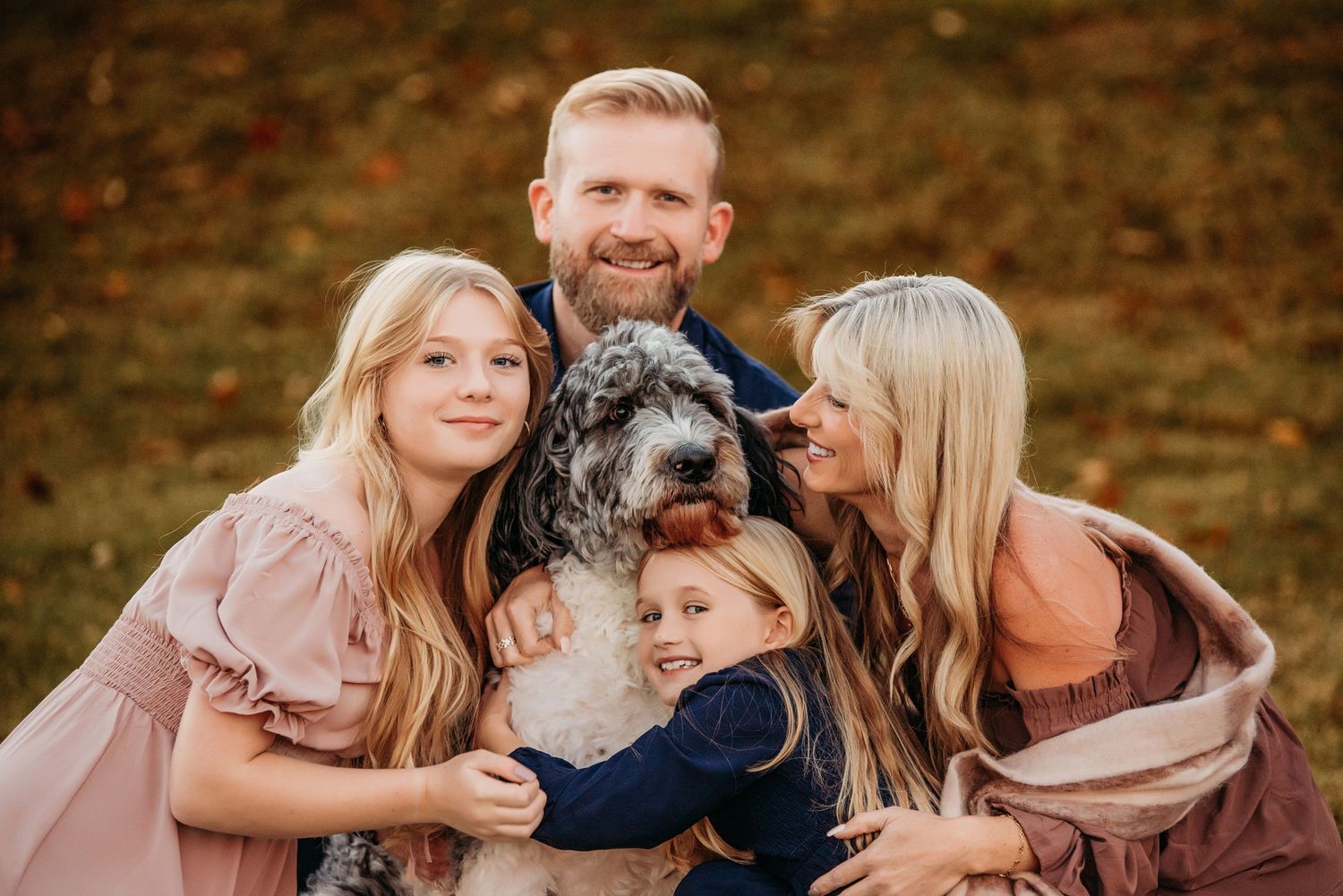 Family of four with two blonde girls and parents smiling outdoors, hugging a fluffy gray dog in autumn setting.