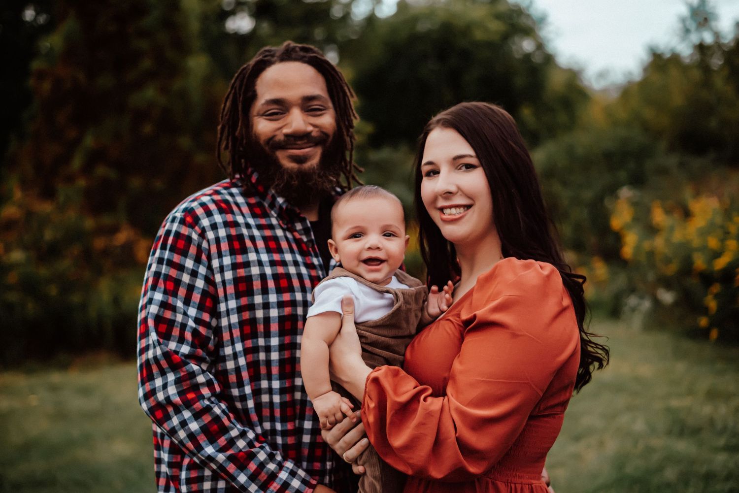 A smiling family of three poses outdoors in fall, featuring a man, woman in orange, and their happy baby.