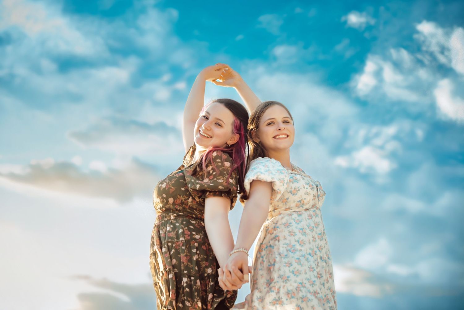 Two young women in floral dresses hold hands and pose together against a bright blue sky with white clouds.