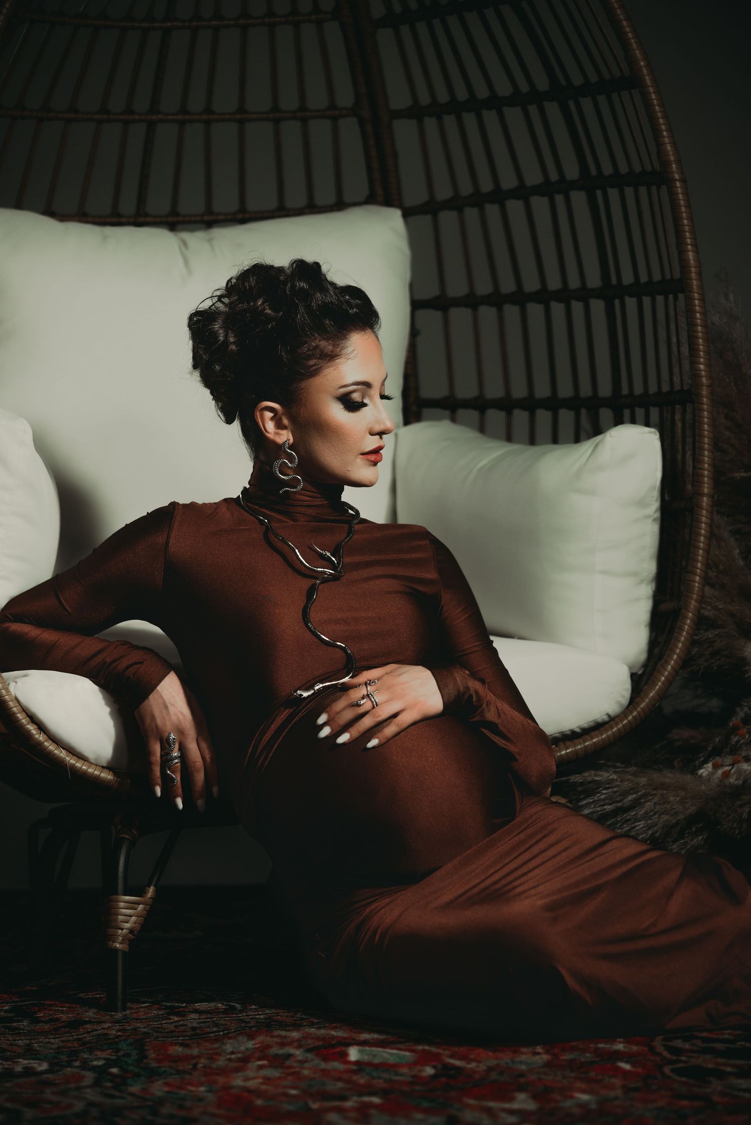 Elegant maternity portrait in burgundy dress against wicker chair and white cushions.