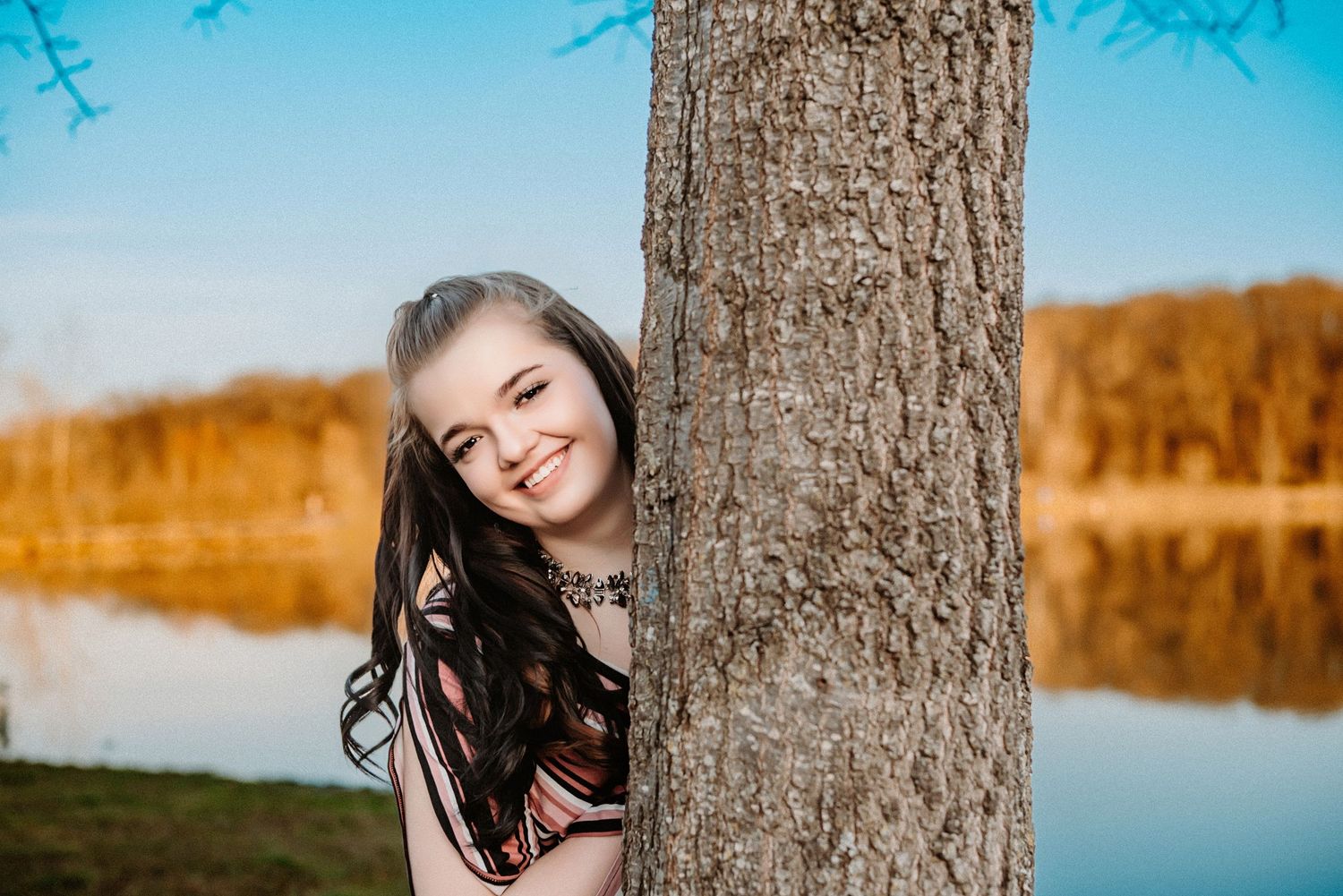 A smiling young woman with long dark hair peeks playfully from behind a tree trunk near a calm lake at sunset.