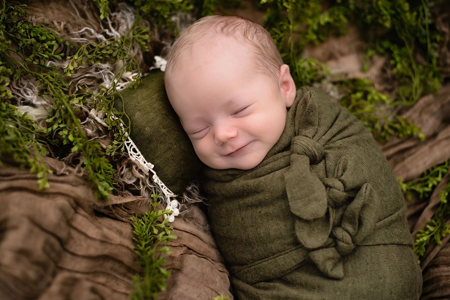 Smiling newborn baby swaddled in green wrap, nestled among moss and white flowers in a nature-themed photo.