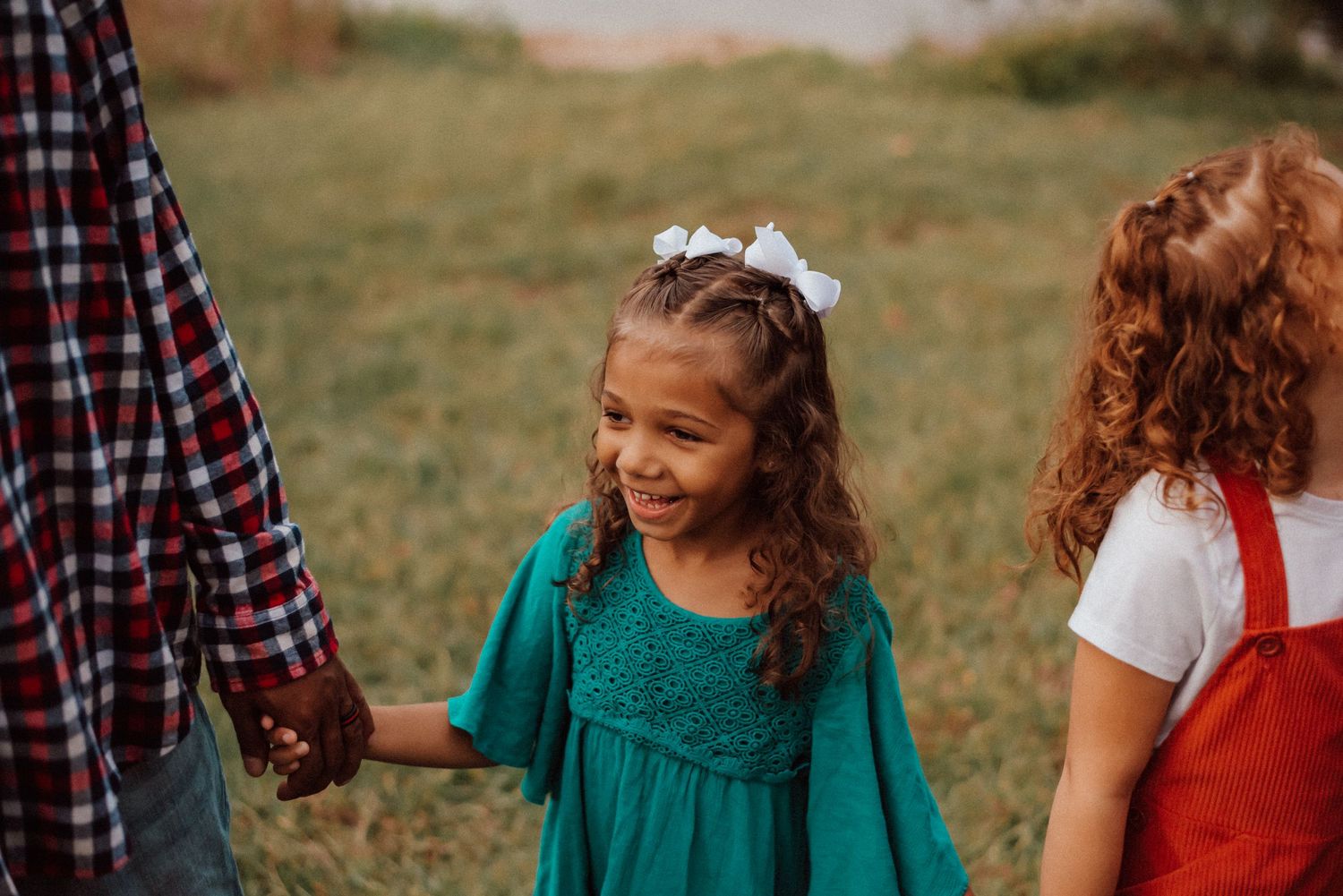 Two young girls holding hands outdoors, one in a teal dress with white bow, another in red overalls.
