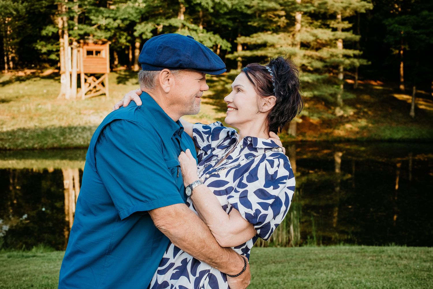 Happy couple embracing and smiling outdoors near a pond surrounded by lush green trees and grass.