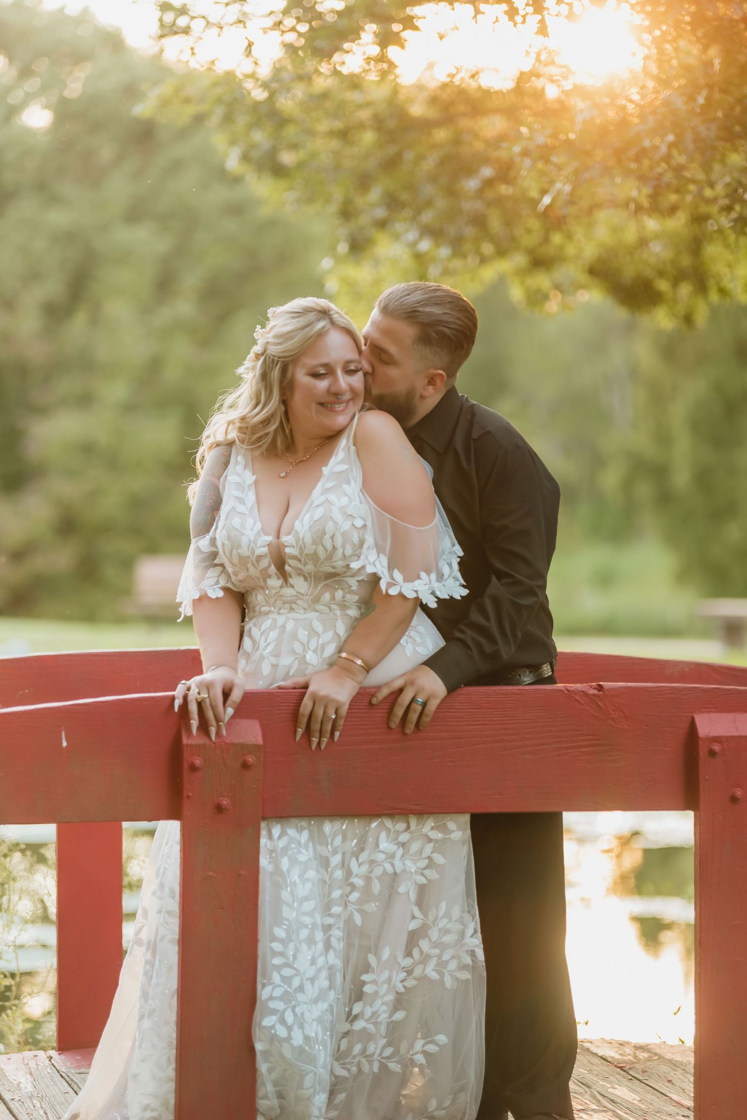 Bride and groom share a romantic moment on a red bridge at golden hour, surrounded by lush greenery.