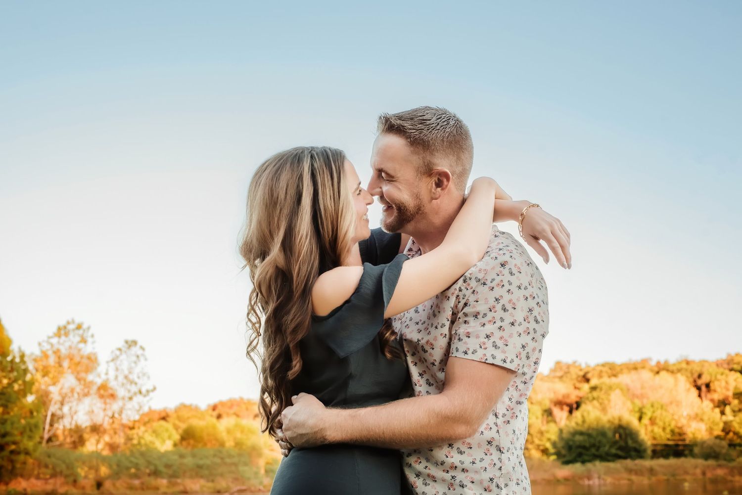 Romantic couple embraces during golden hour sunset by a lake with autumn foliage in background.