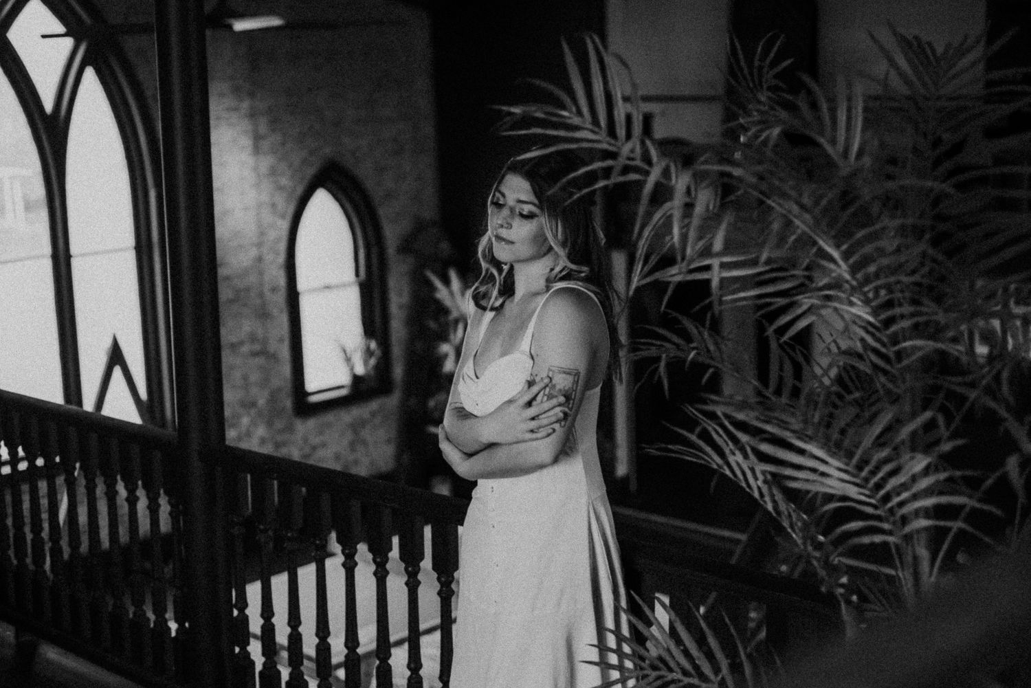 Black and white photo of a bride in an elegant dress standing near arched windows and tropical plants.