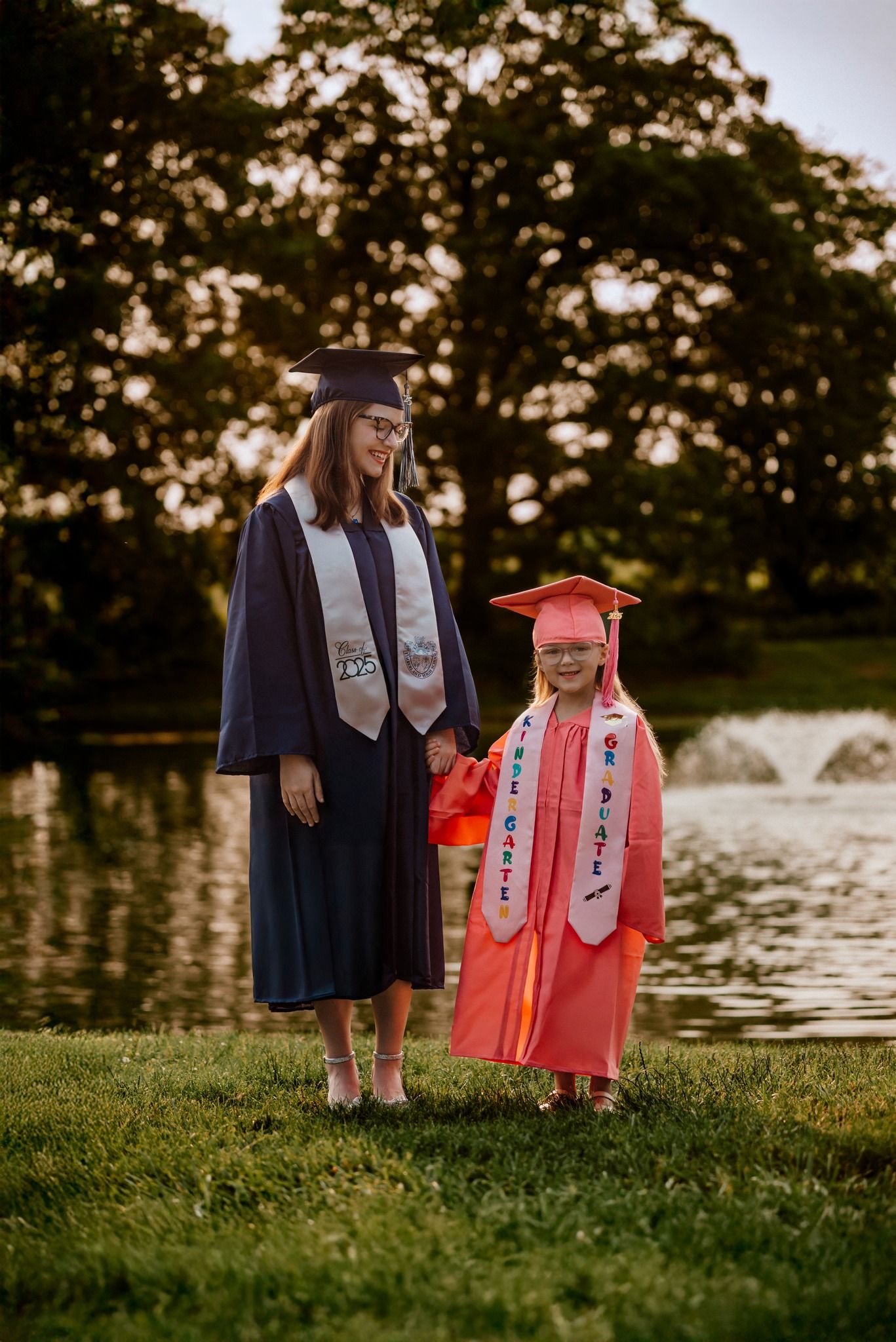 Graduates in traditional caps and gowns standing by a lake at sunset.