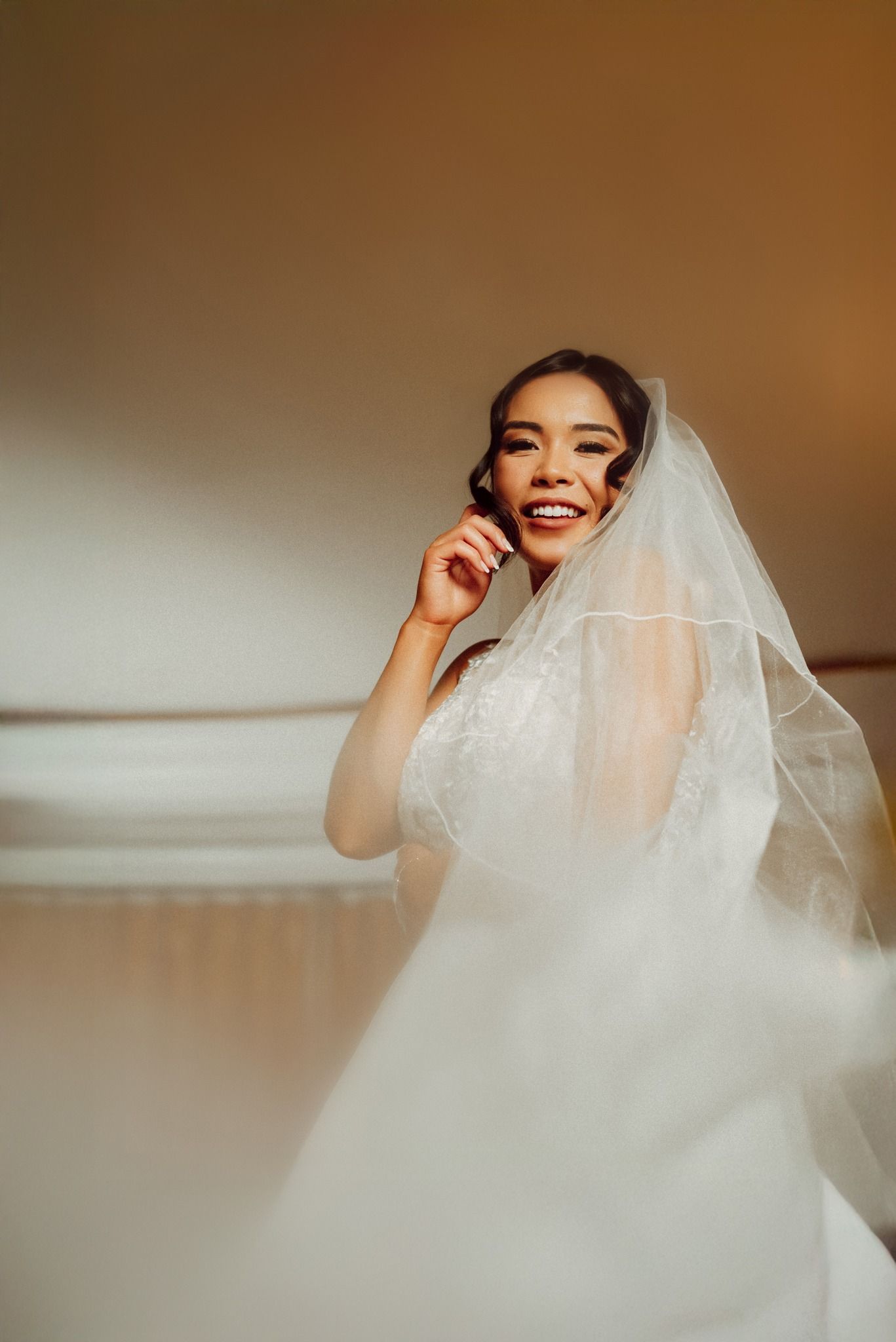 Bride wearing white veil laughing and smiling in warm lighting during wedding preparations.