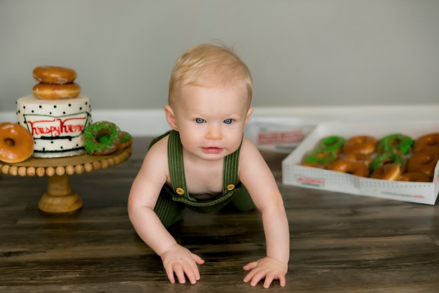 Baby in green overalls crawls on wooden table near donuts and party decorations.