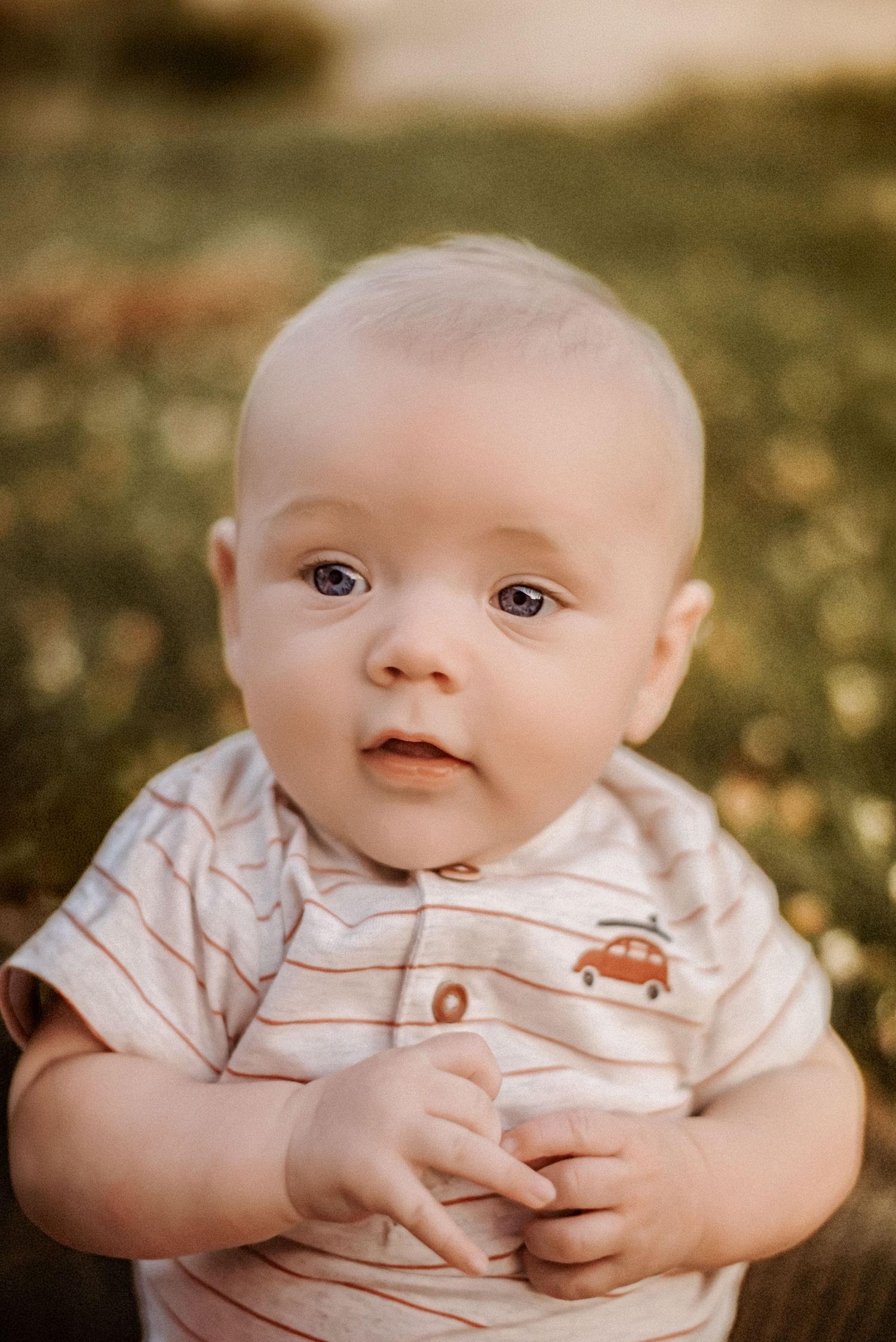 Adorable baby in striped onesie with car print sits outdoors in golden light, looking curiously at camera.
