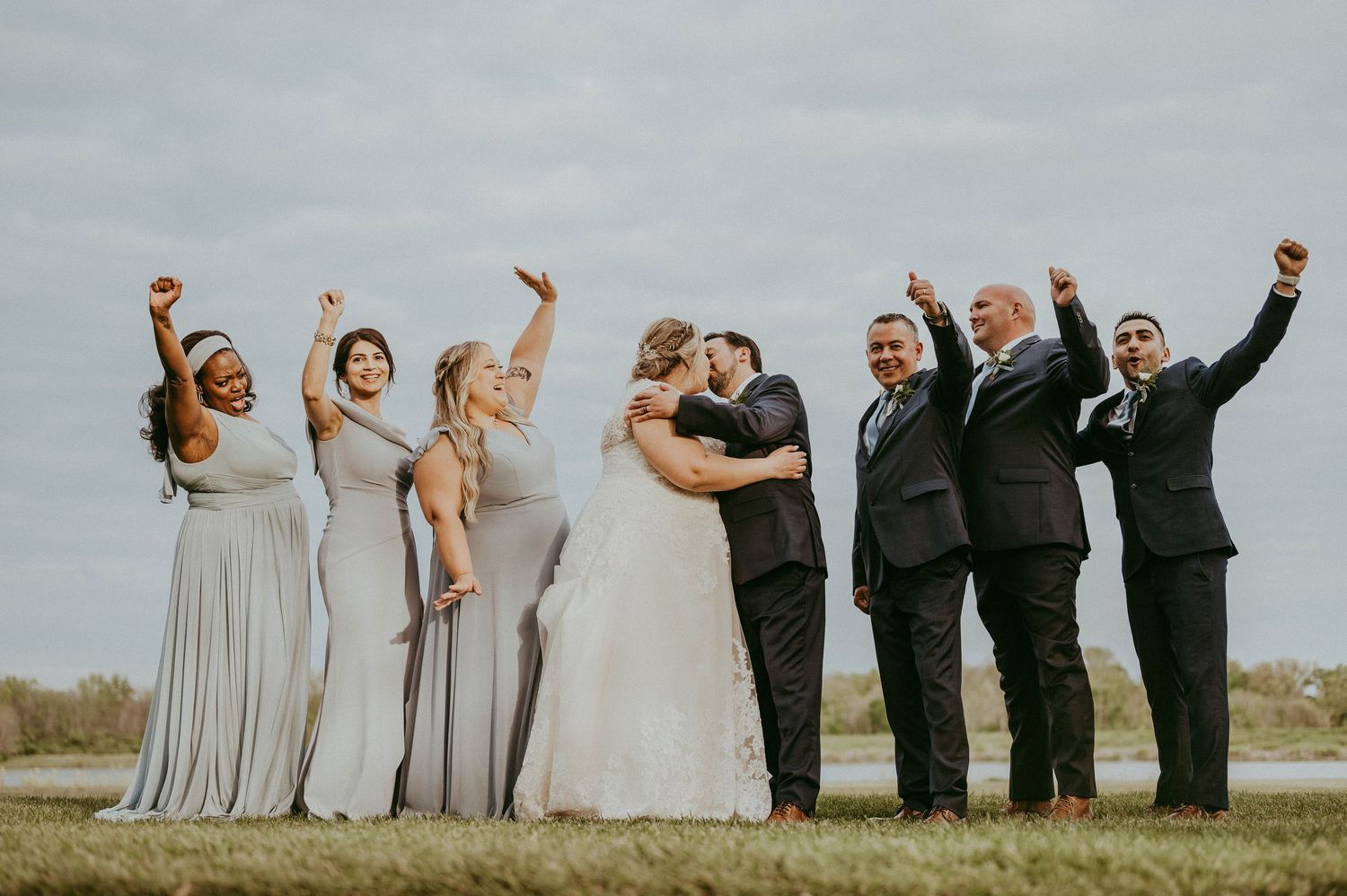 Wedding party celebrating with raised arms as bride and groom kiss outdoors, bridesmaids in gray gowns.