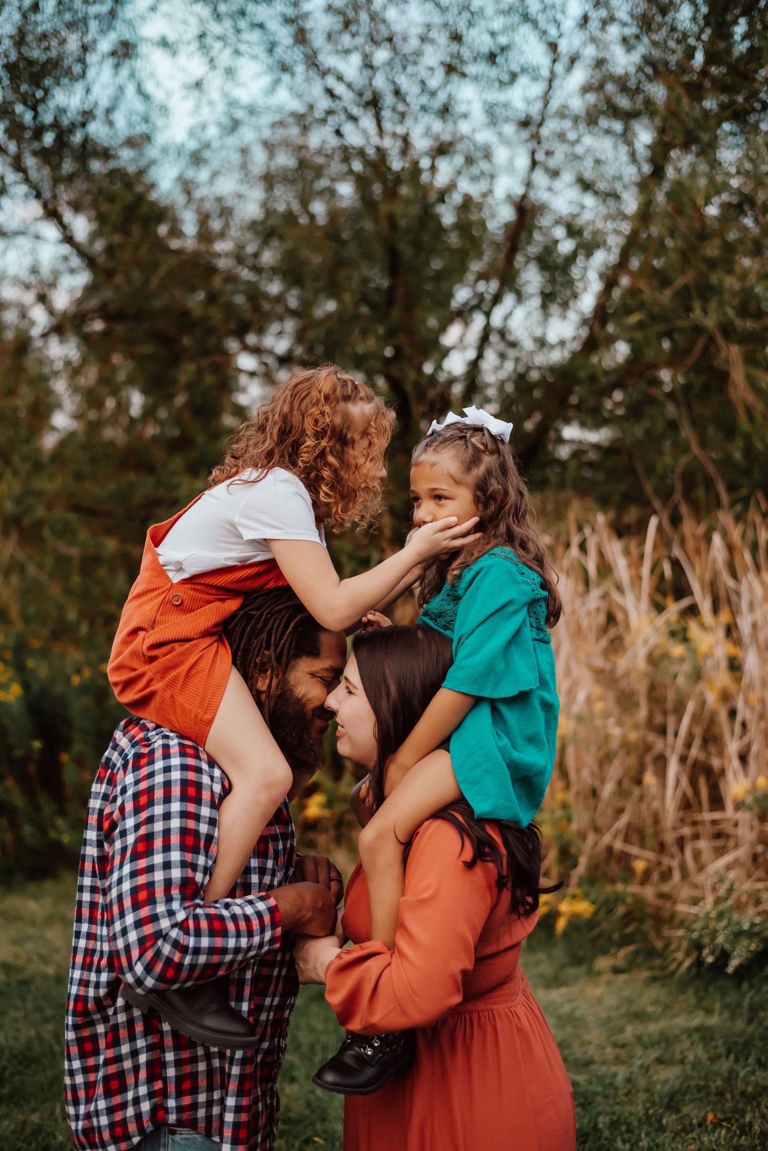 A joyful family of four outdoors, with two children sitting on parents shoulders, smiling and playing in a green park.