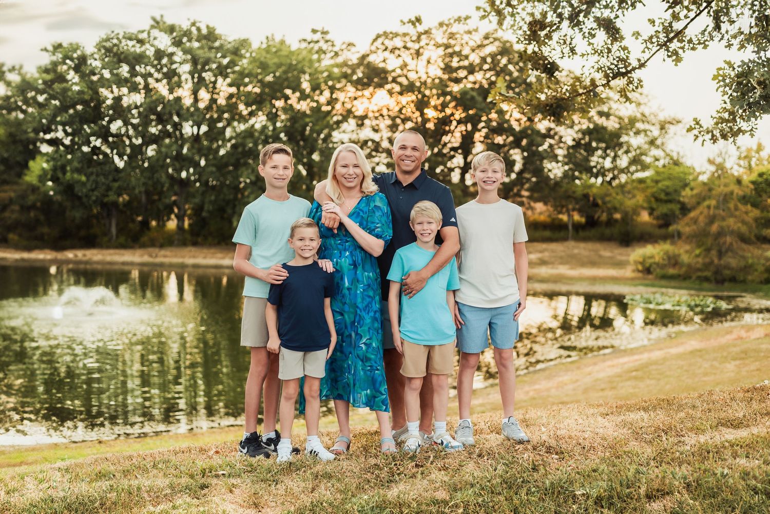 Family of six posing outdoors near a pond at golden hour, surrounded by lush green trees.