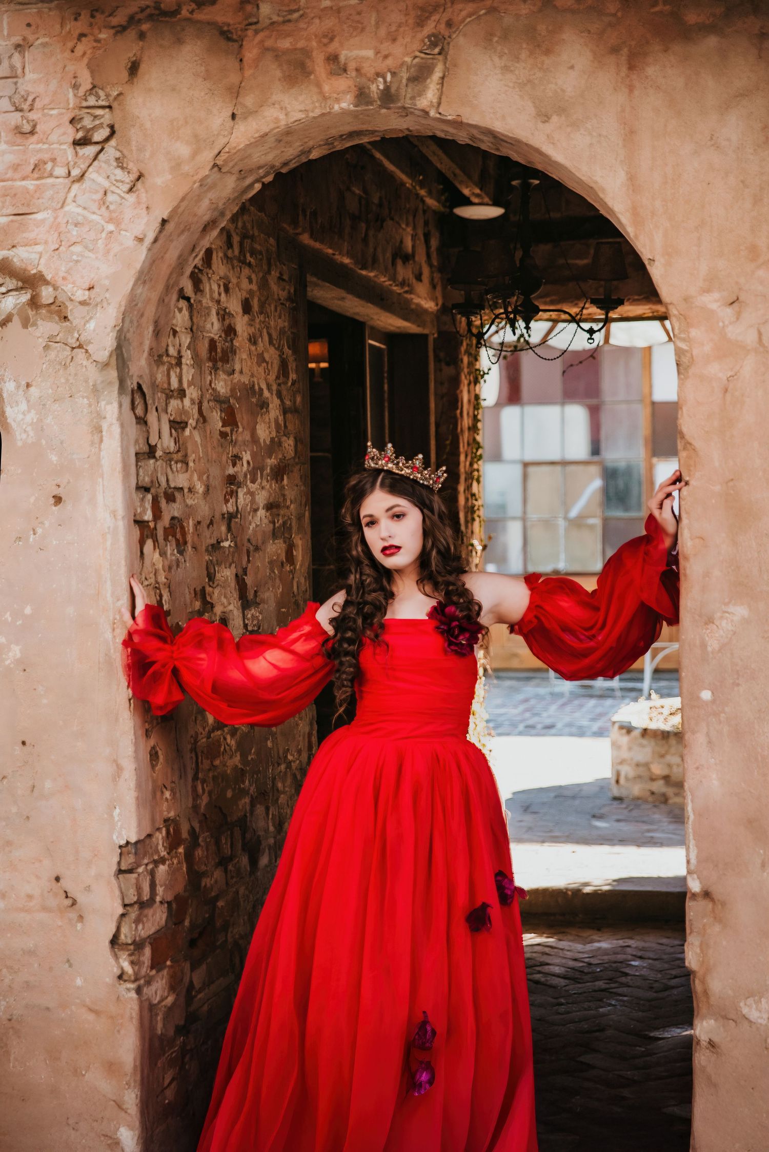 Young woman in red ballgown and crown poses in stone archway doorway in elegant royal-themed portrait.