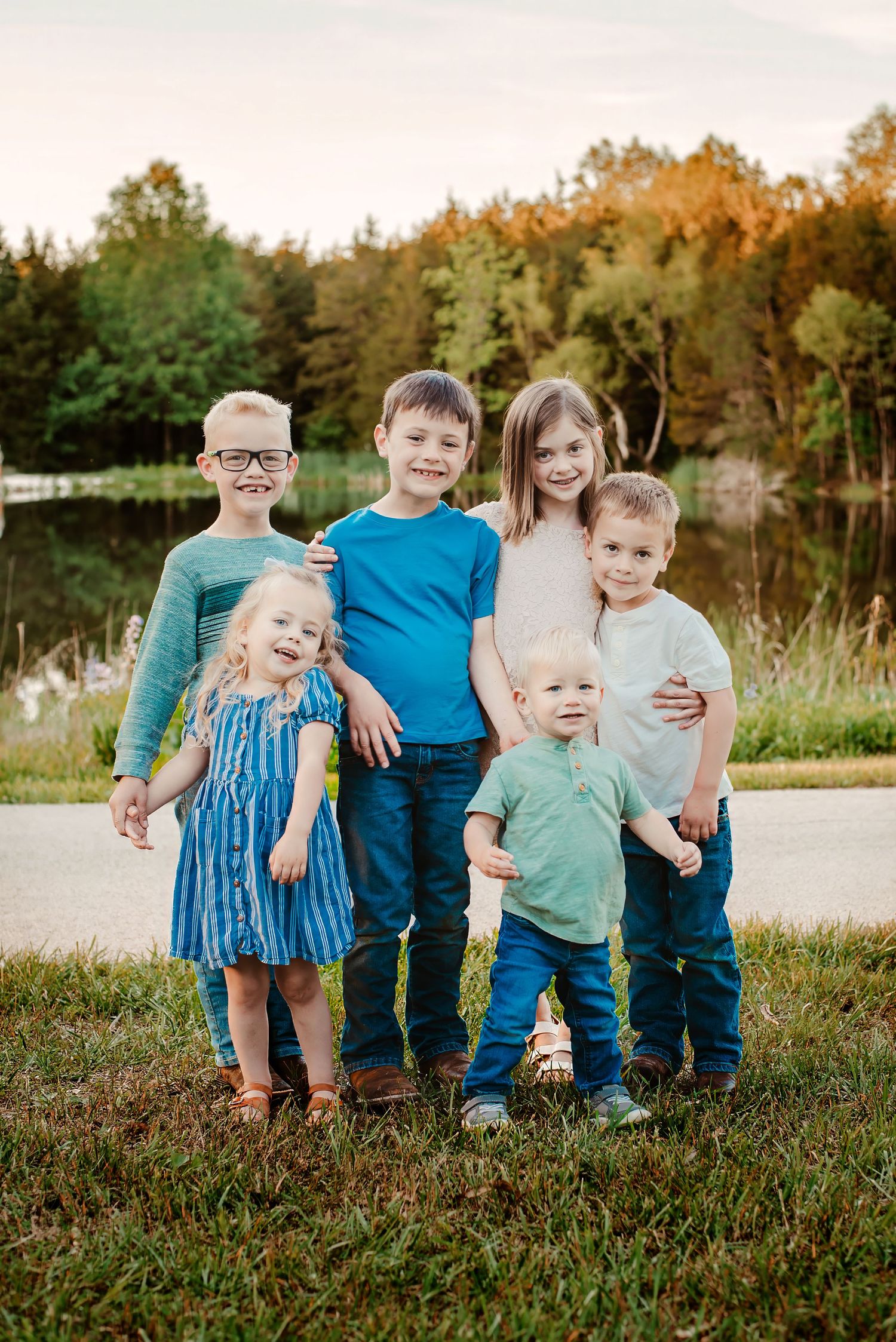 Six young children smiling and posing together outdoors near a pond with autumn trees in the background.