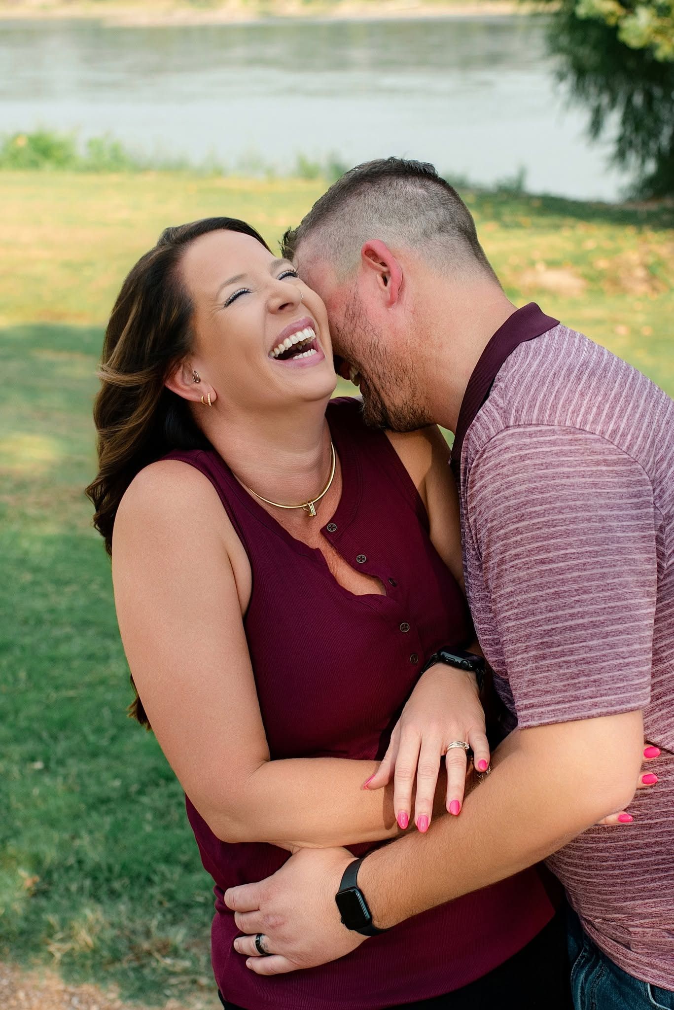 A joyful couple embraces outdoors near a river, woman laughing as man kisses her cheek, wearing matching burgundy outfits.