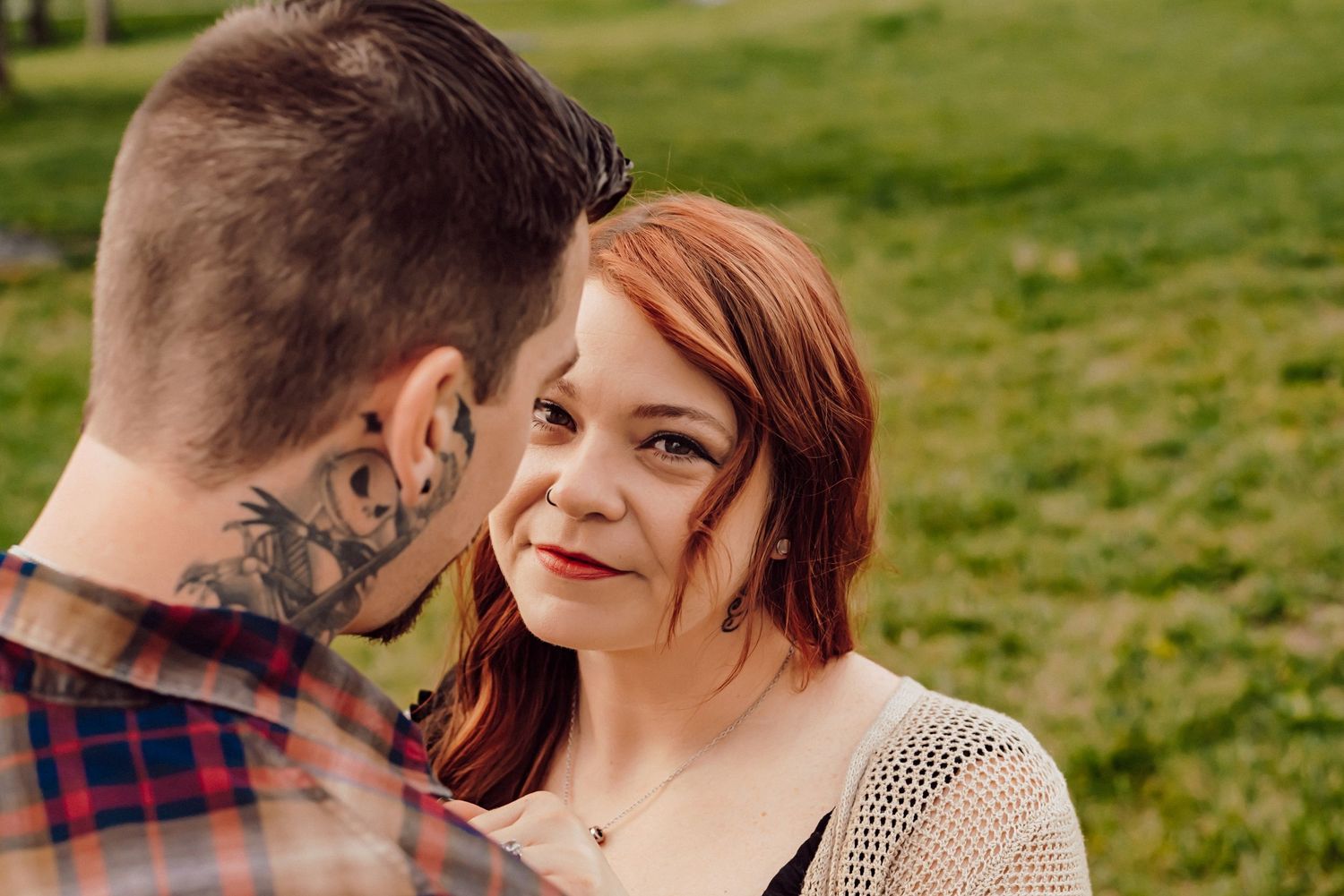 A redhead woman gazes at a tattooed man outdoors in a green field during a romantic couples portrait session.