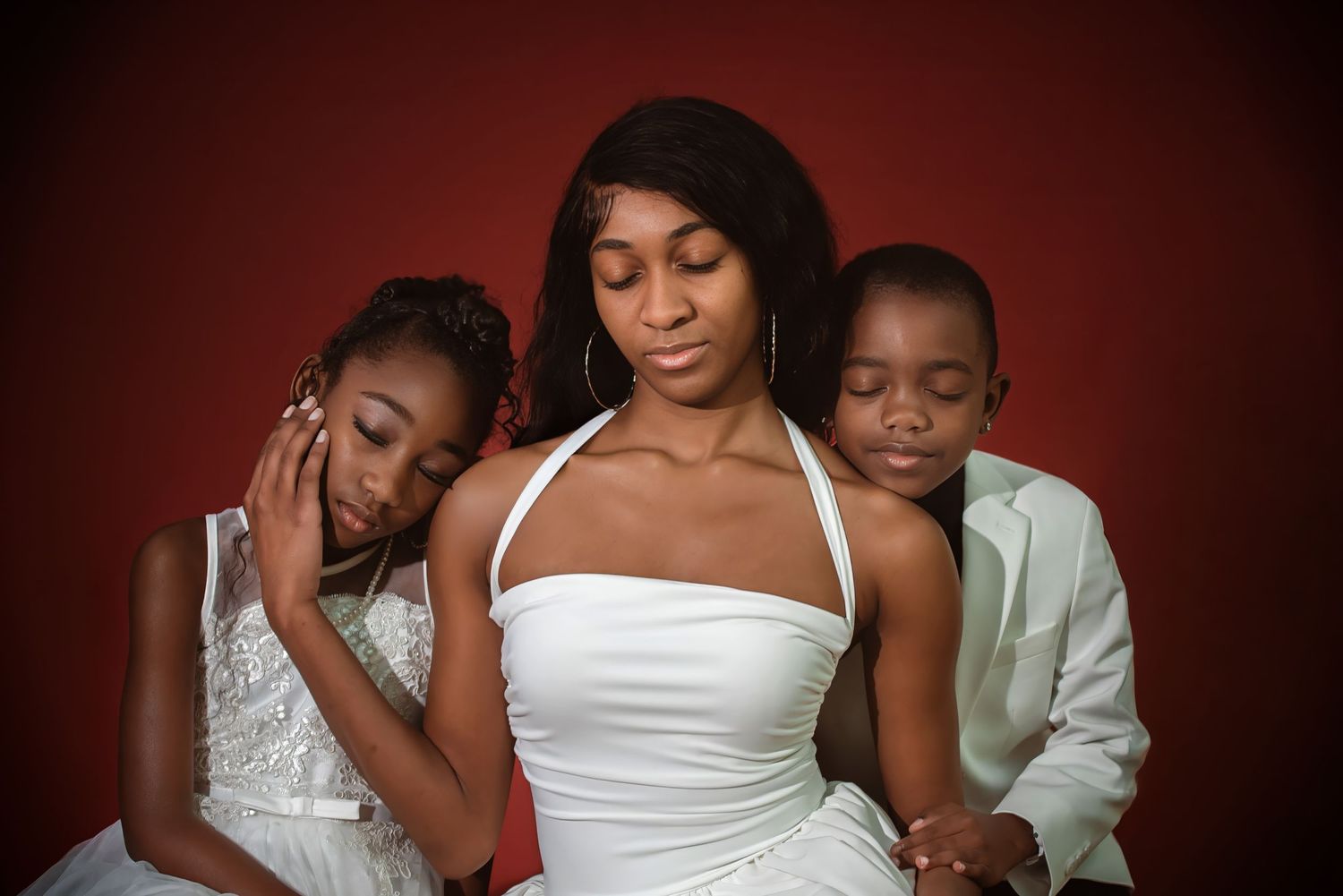 A woman in a white dress sits between two children in formal wear against a red background.