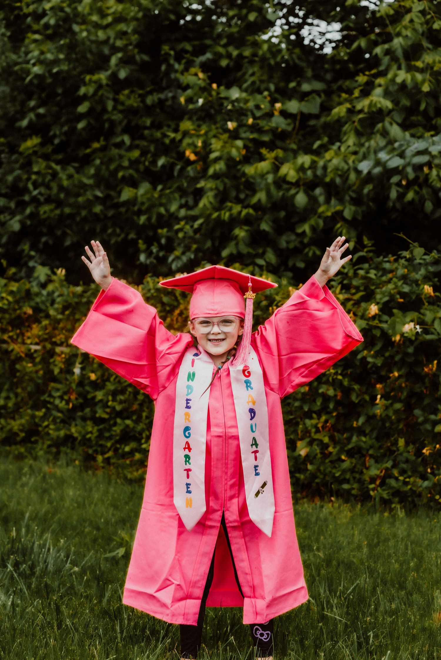 A graduate in bright pink regalia throws their arms up in celebration outdoors.
