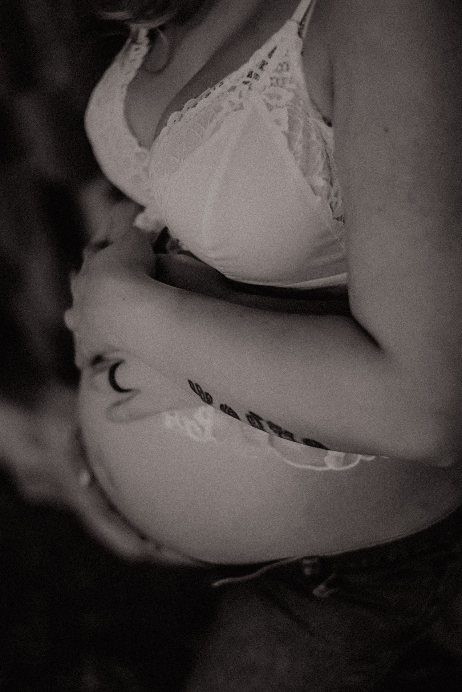 A black and white artistic photograph showing a pregnant belly with a small moon tattoo, wearing white lace lingerie.