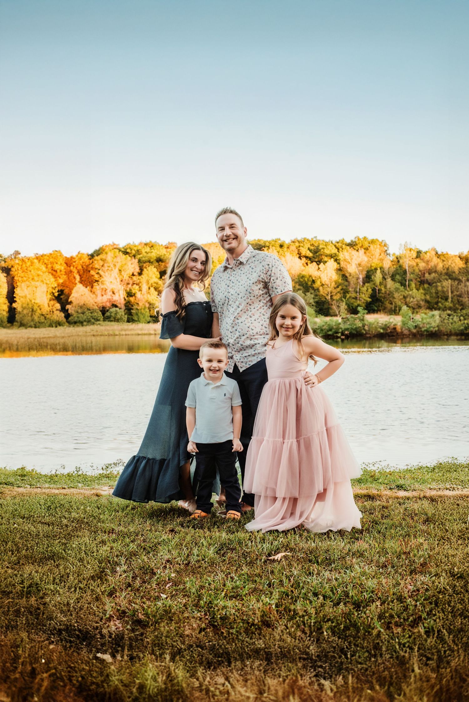 Family in coordinated outfits poses by lake during golden hour with fall foliage in background.