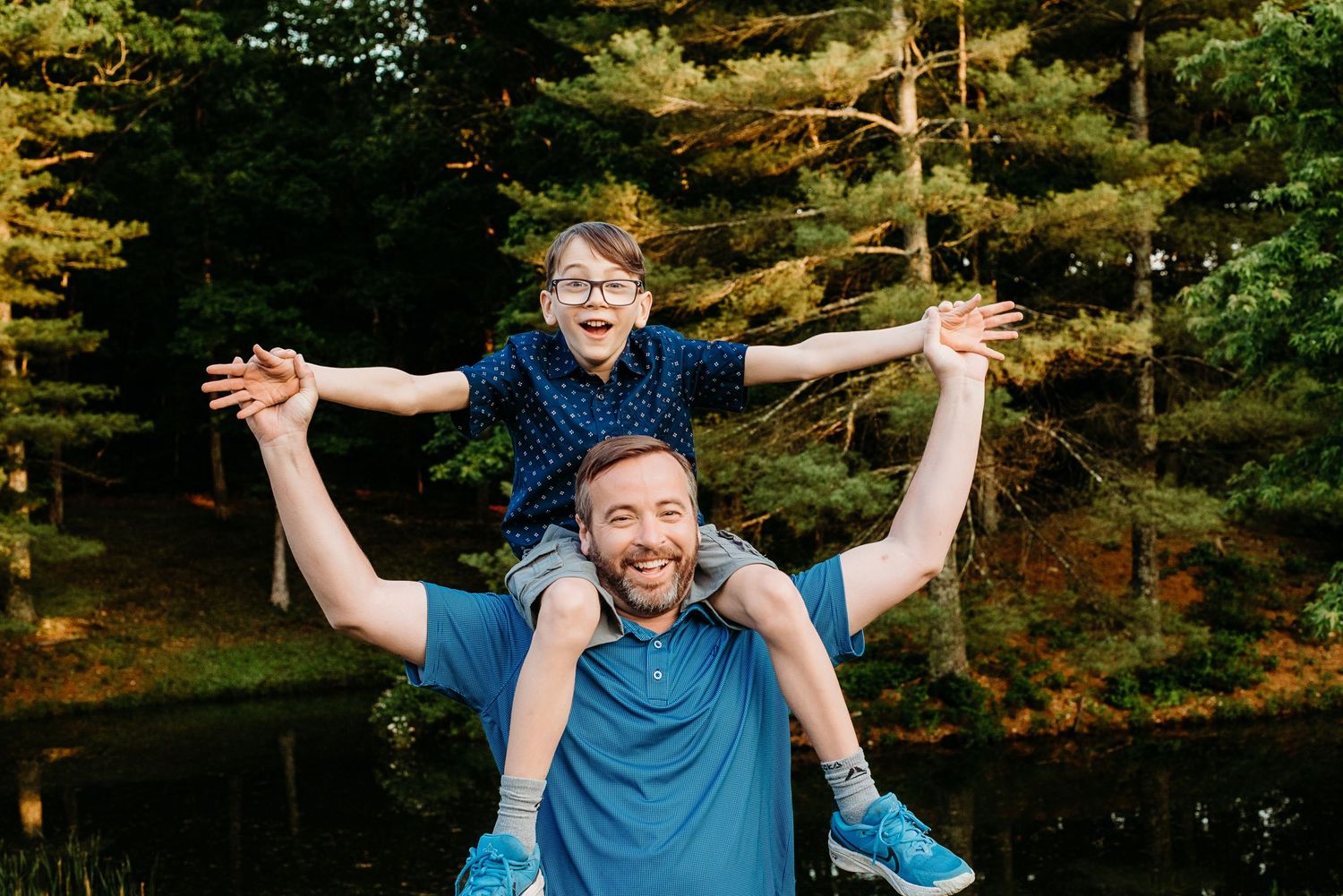 A joyful father carries his excited young son on his shoulders outdoors near a pond surrounded by autumn trees.